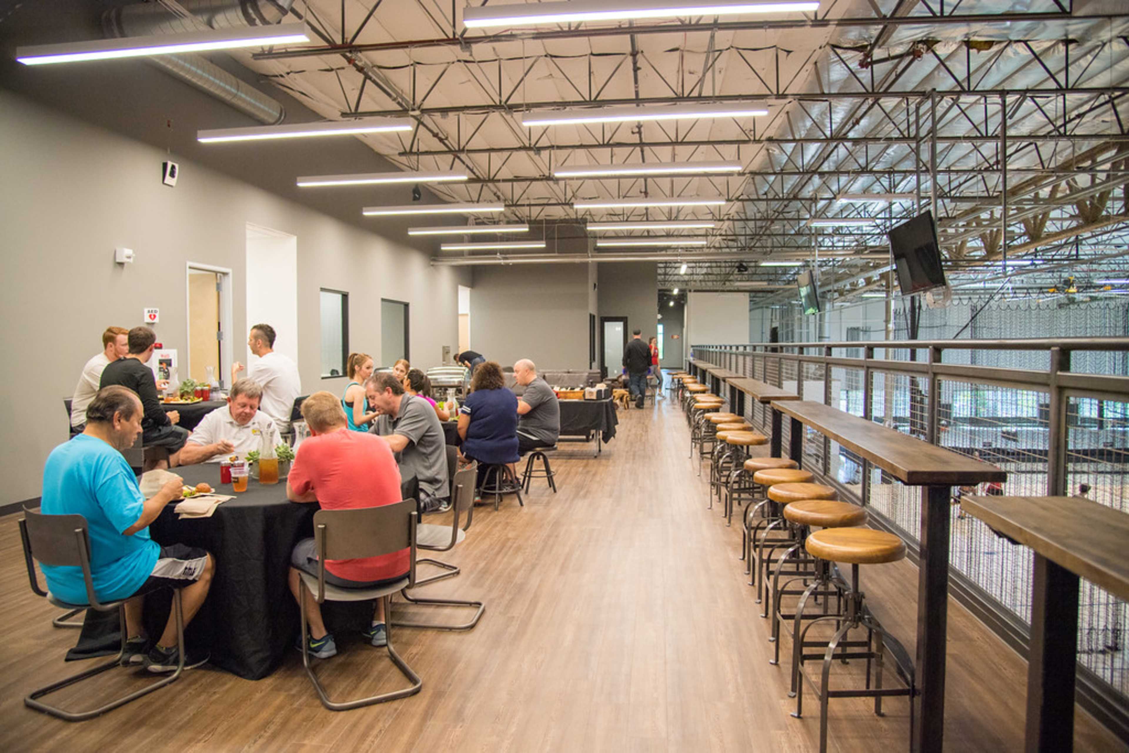 A group of people sits at tables and high-top seating in a spacious indoor venue, sharing a meal and conversation.