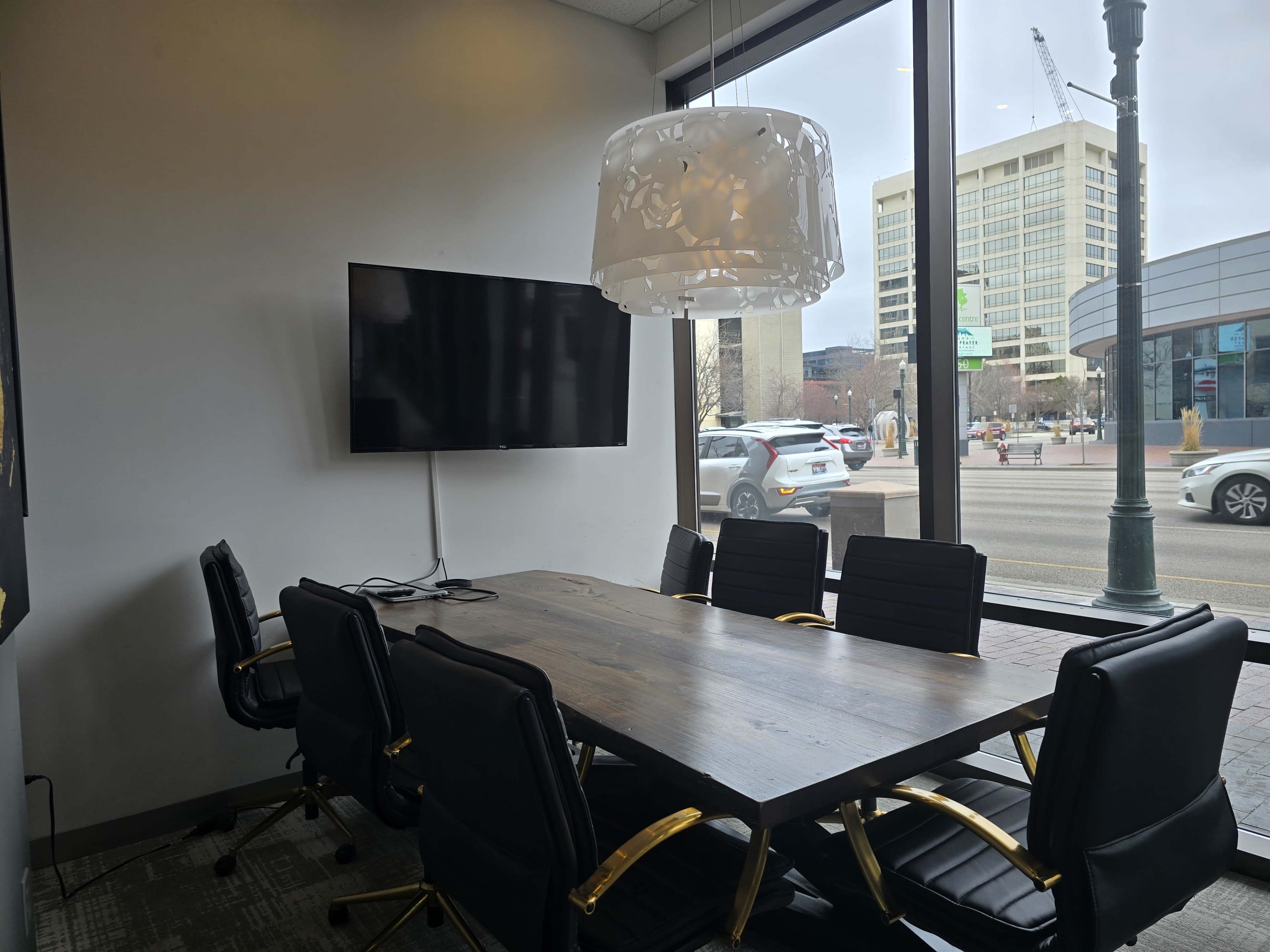 A conference room features a wooden table surrounded by black chairs, with a mounted television on the wall and large windows overlooking a city street.
