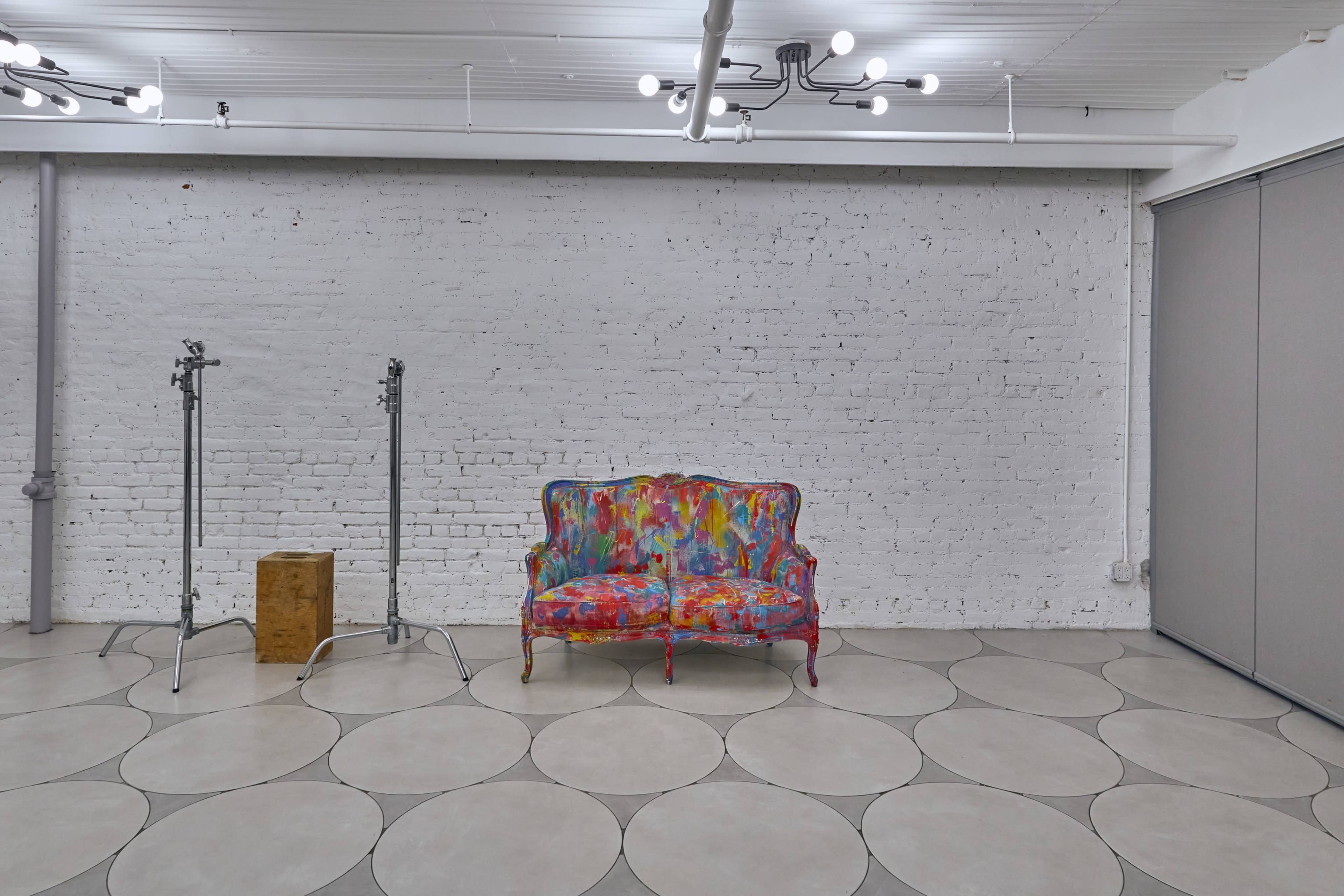 A colorful couch sits in a minimalist room with a white brick wall and light fixtures above.