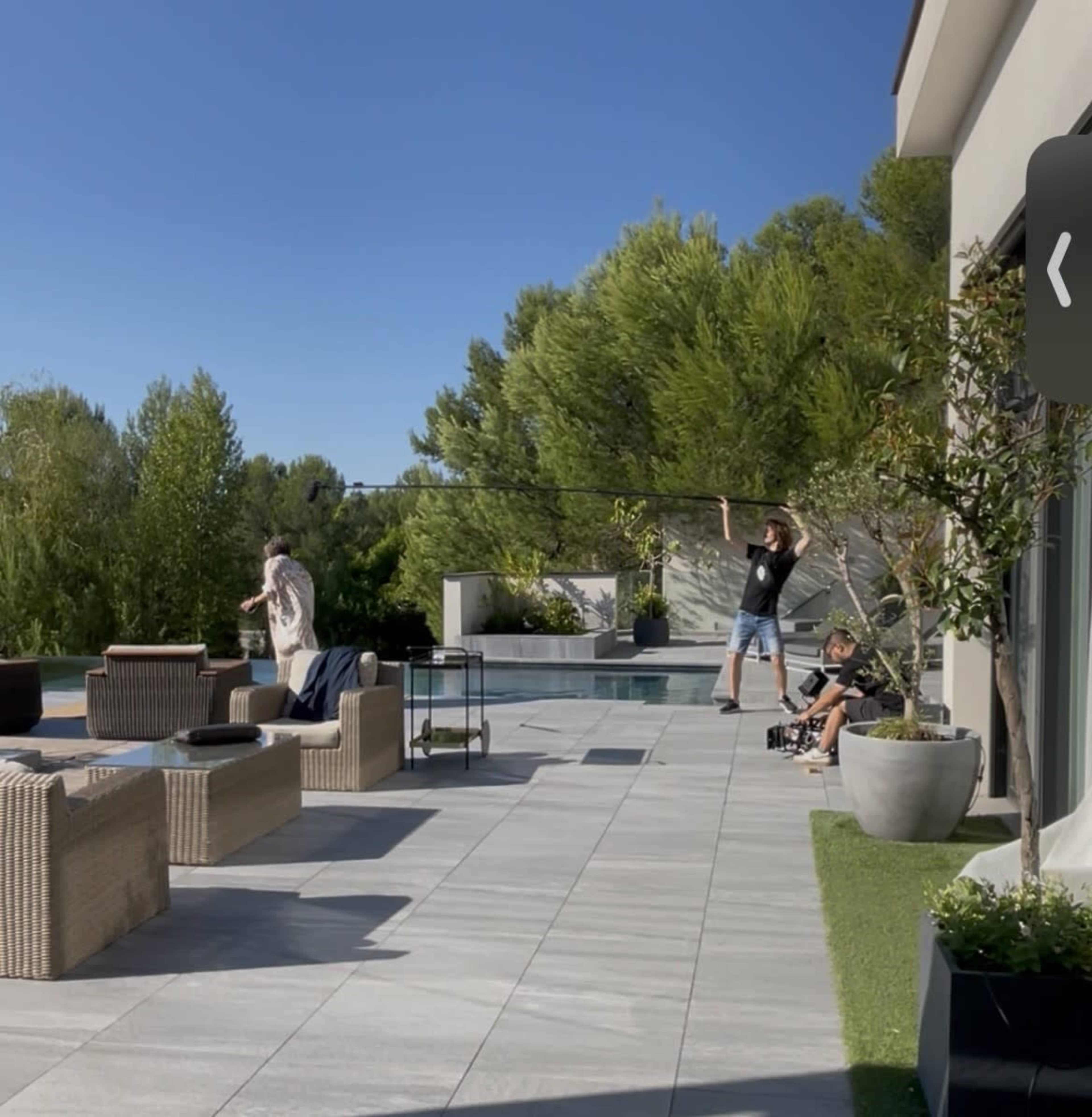 A film crew is set up on a patio with outdoor furniture, capturing a scene near a pool surrounded by greenery.