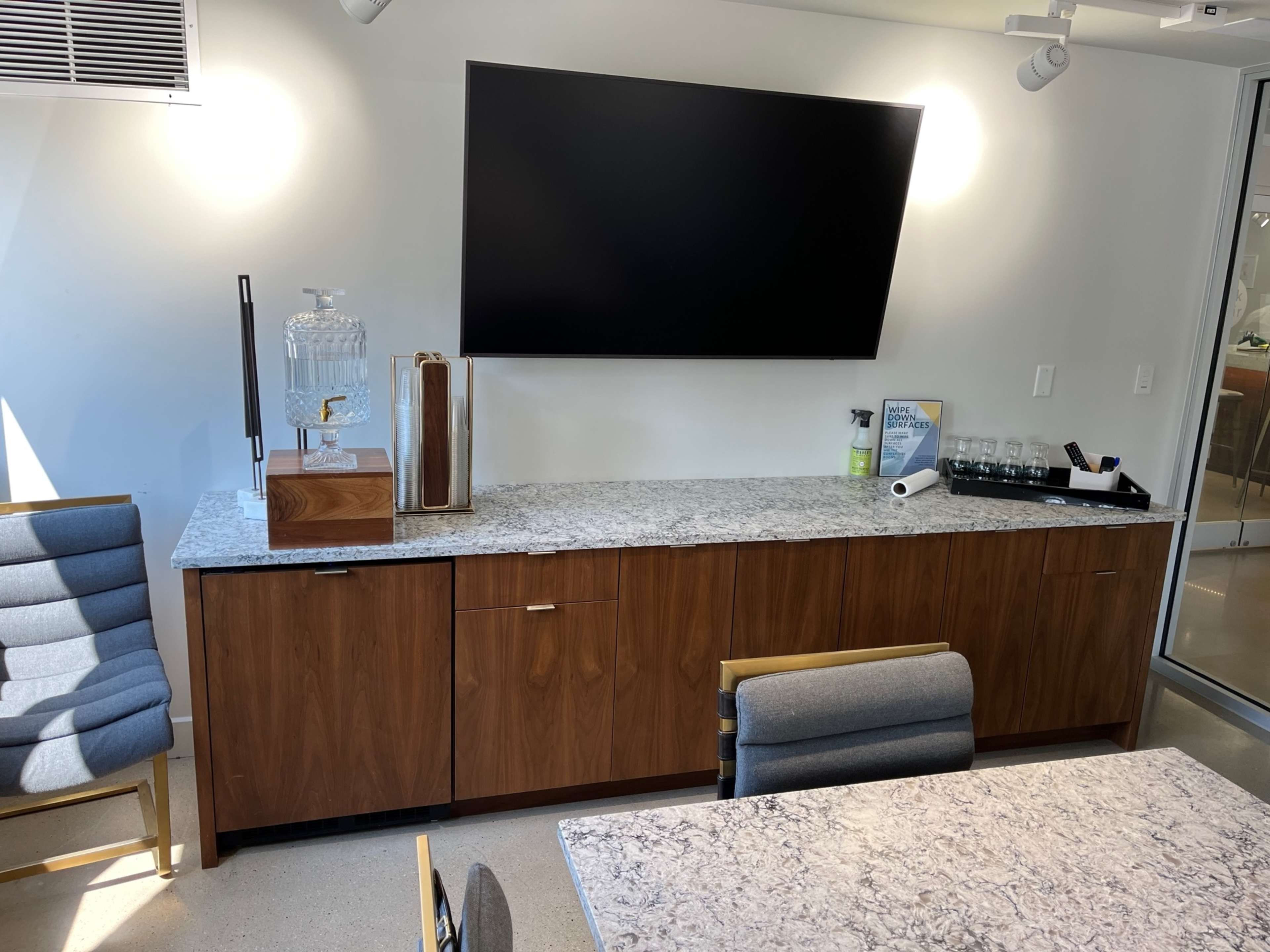 A modern conference room with a wooden cabinet below a wall-mounted television, a stone countertop, and a seating area featuring a gray chair.