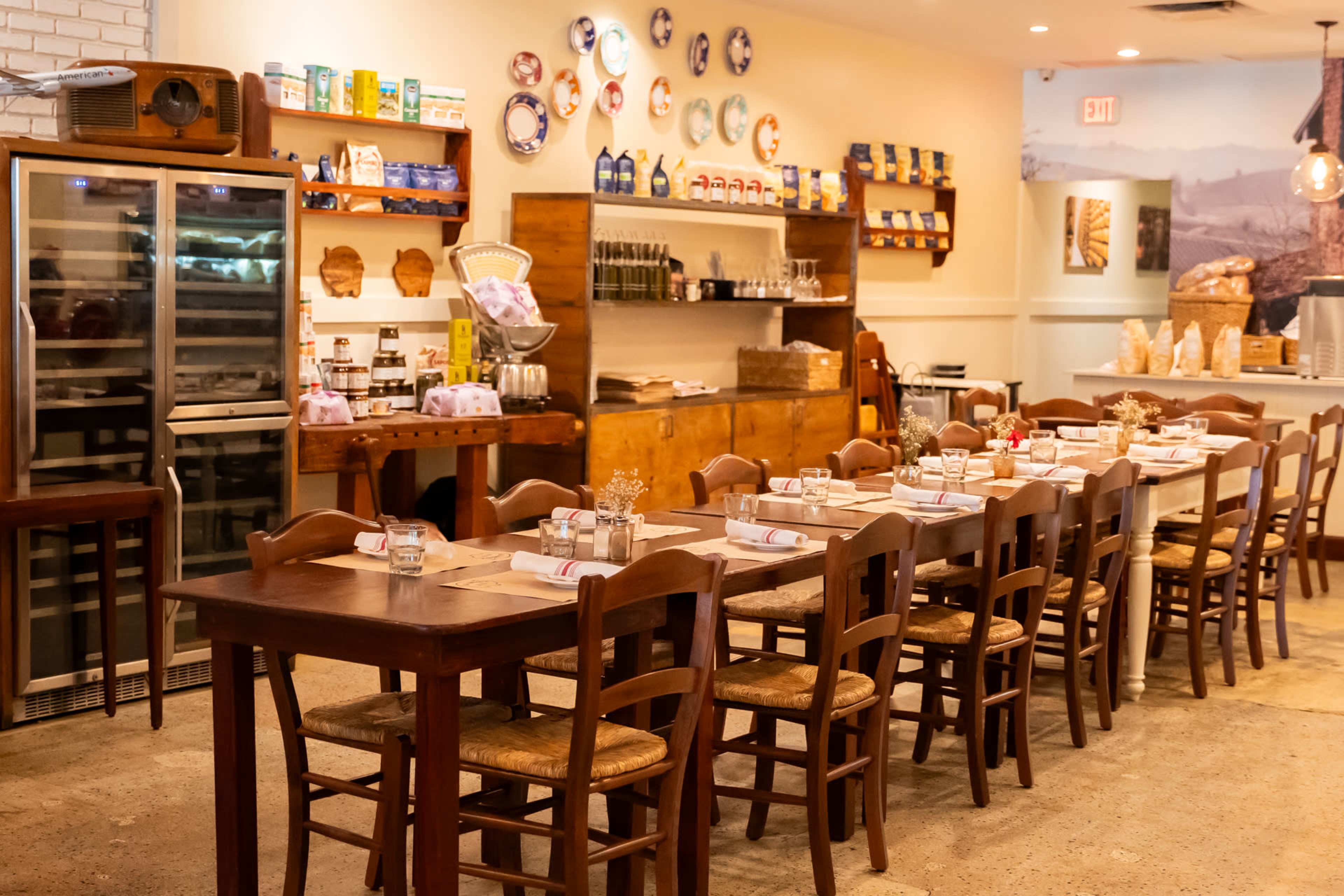 The image shows a cozy restaurant interior with wooden tables and chairs arranged neatly, featuring shelves with various utensils and food products on the walls.