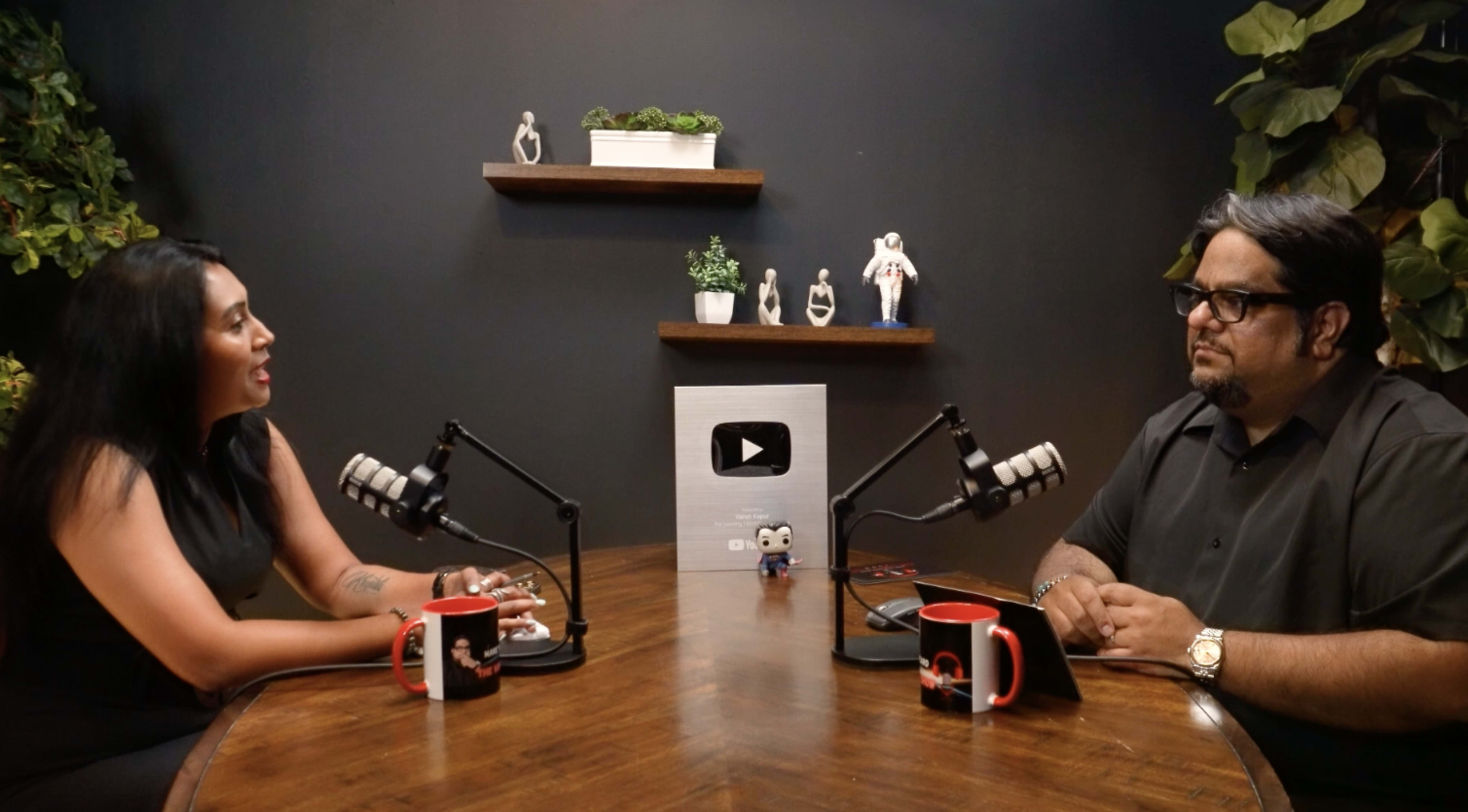 A woman and a man are seated at a wooden table with microphones, facing each other in front of a black wall decorated with shelves displaying figurines and plants.