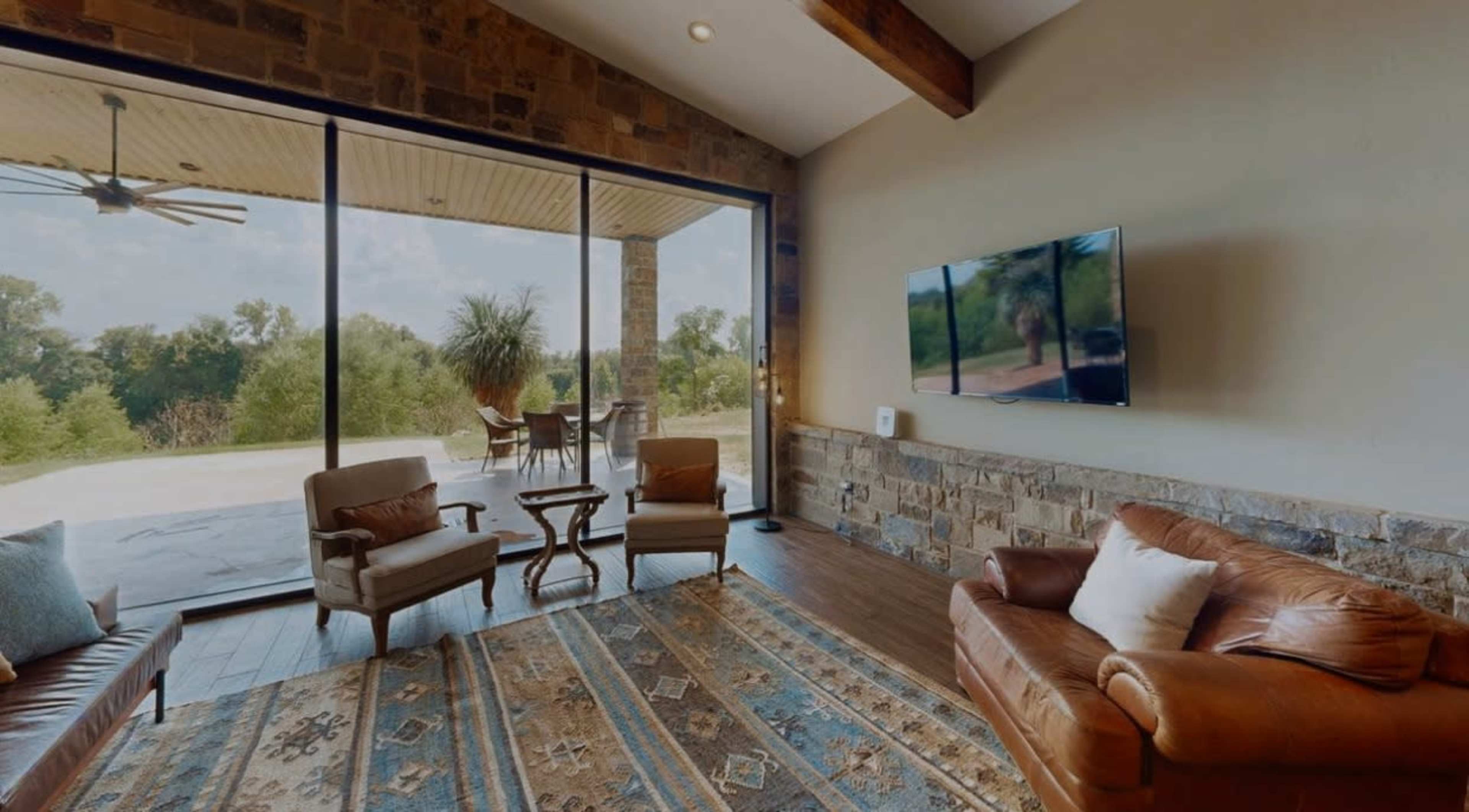 The image shows a living room with a stone wall, two chairs, a brown leather sofa, a coffee table, and a large window overlooking an outdoor patio and greenery.