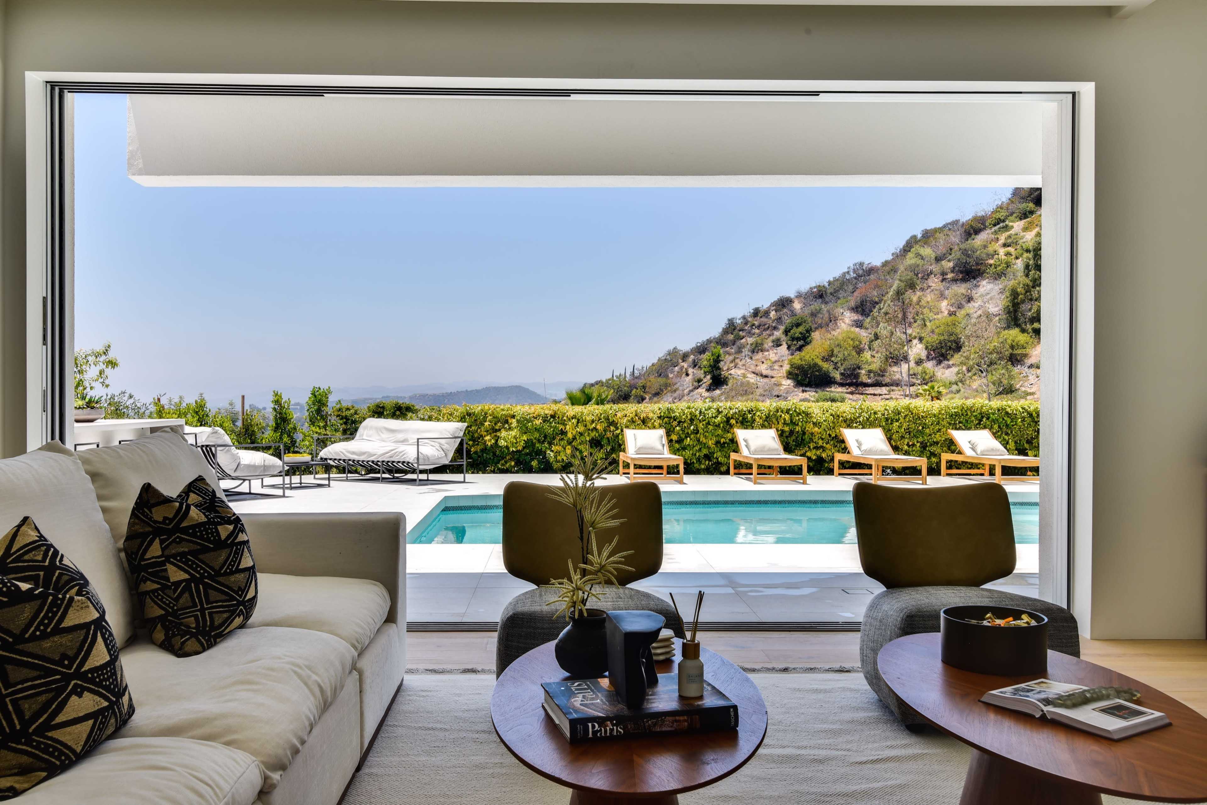 A modern living room with a large opening to a pool area, featuring a white sofa, coffee tables, and a view of mountains and lounge chairs.