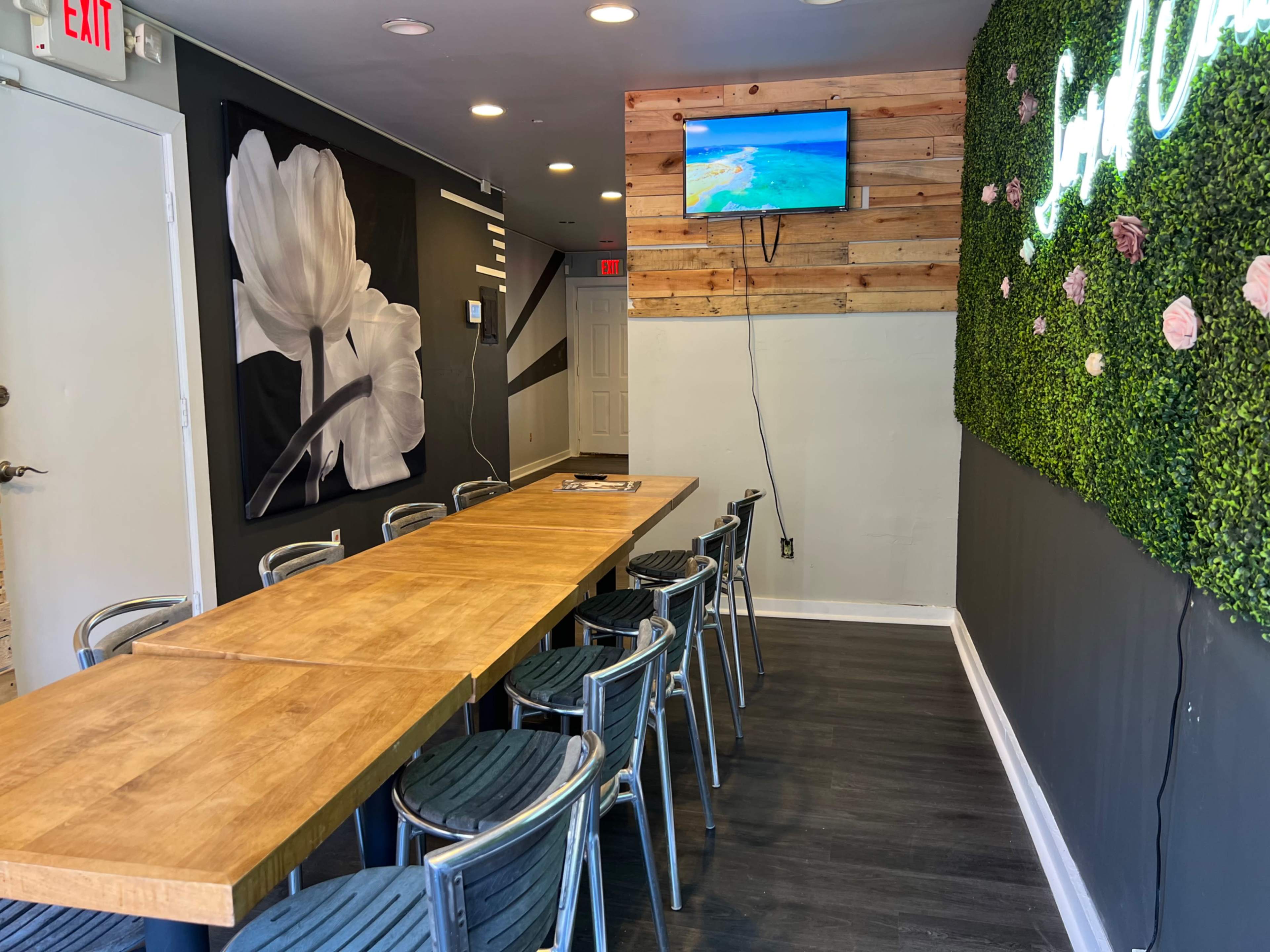 A long wooden table with metal chairs is positioned in a corridor with a floral wall mural and a television screen displaying a beach scene.