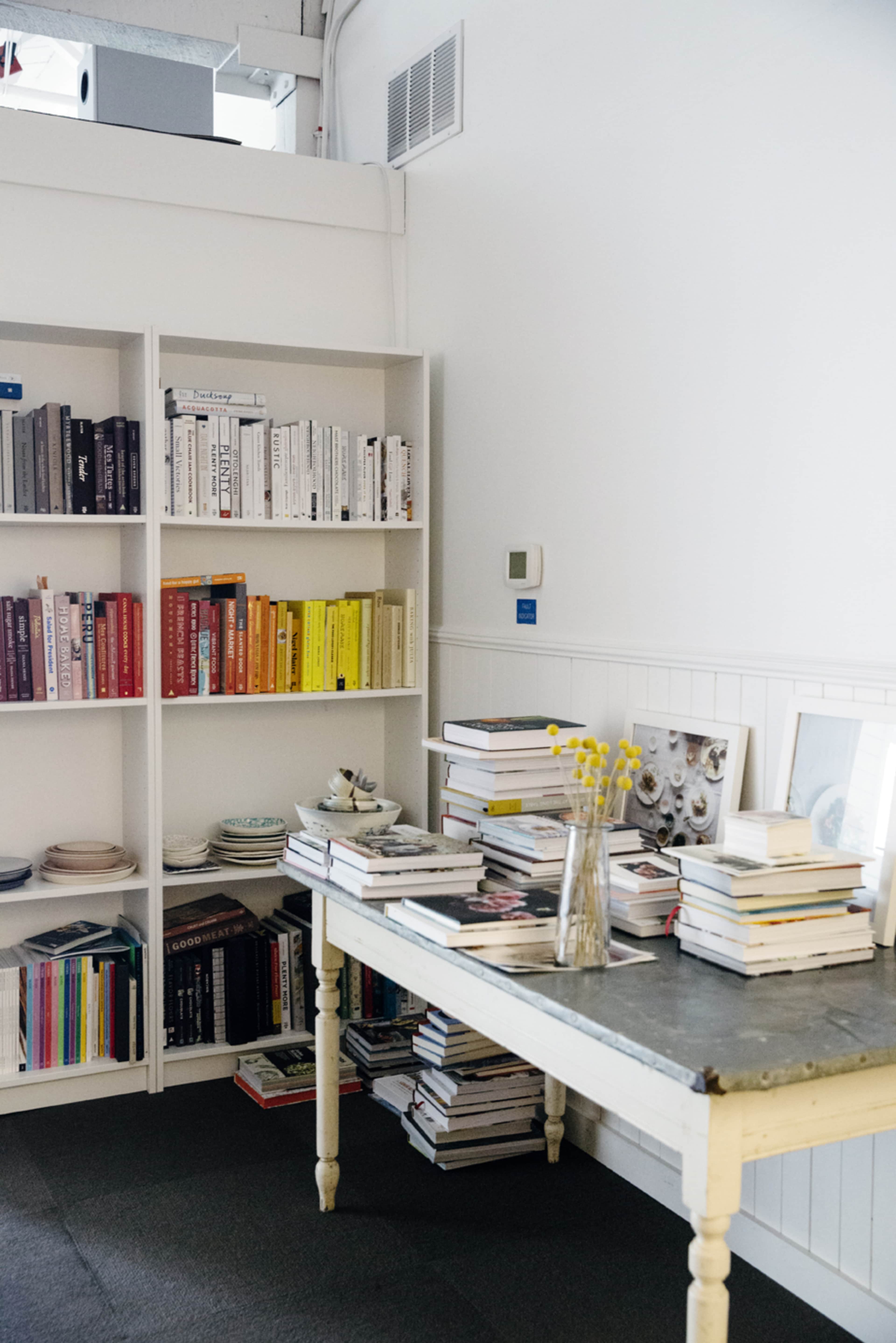 A white-walled room features a bookshelf filled with neatly arranged books, a table stacked with more books, and a vase of yellow flowers.