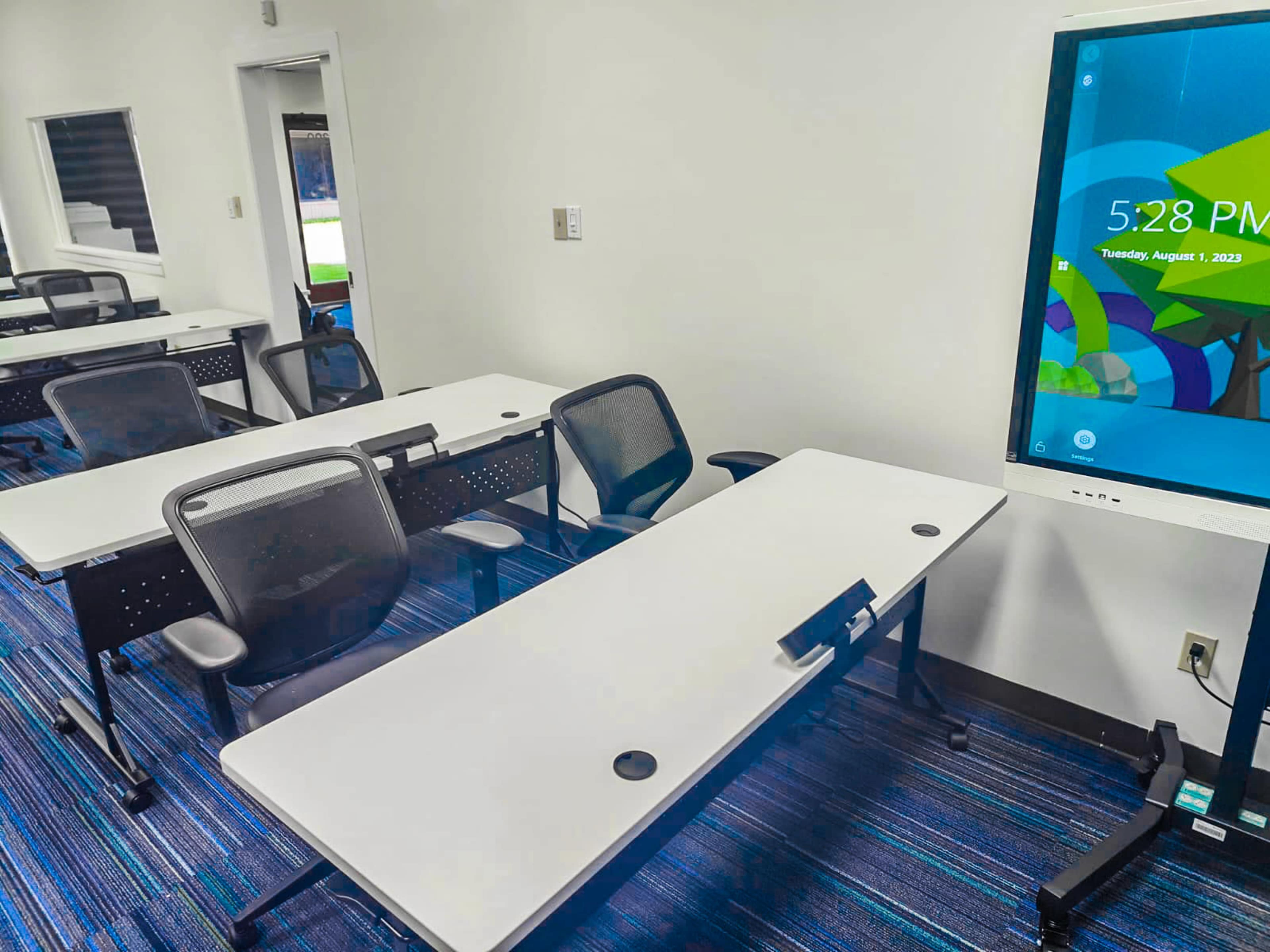 A classroom with several empty desks and a digital screen displaying the date and time.