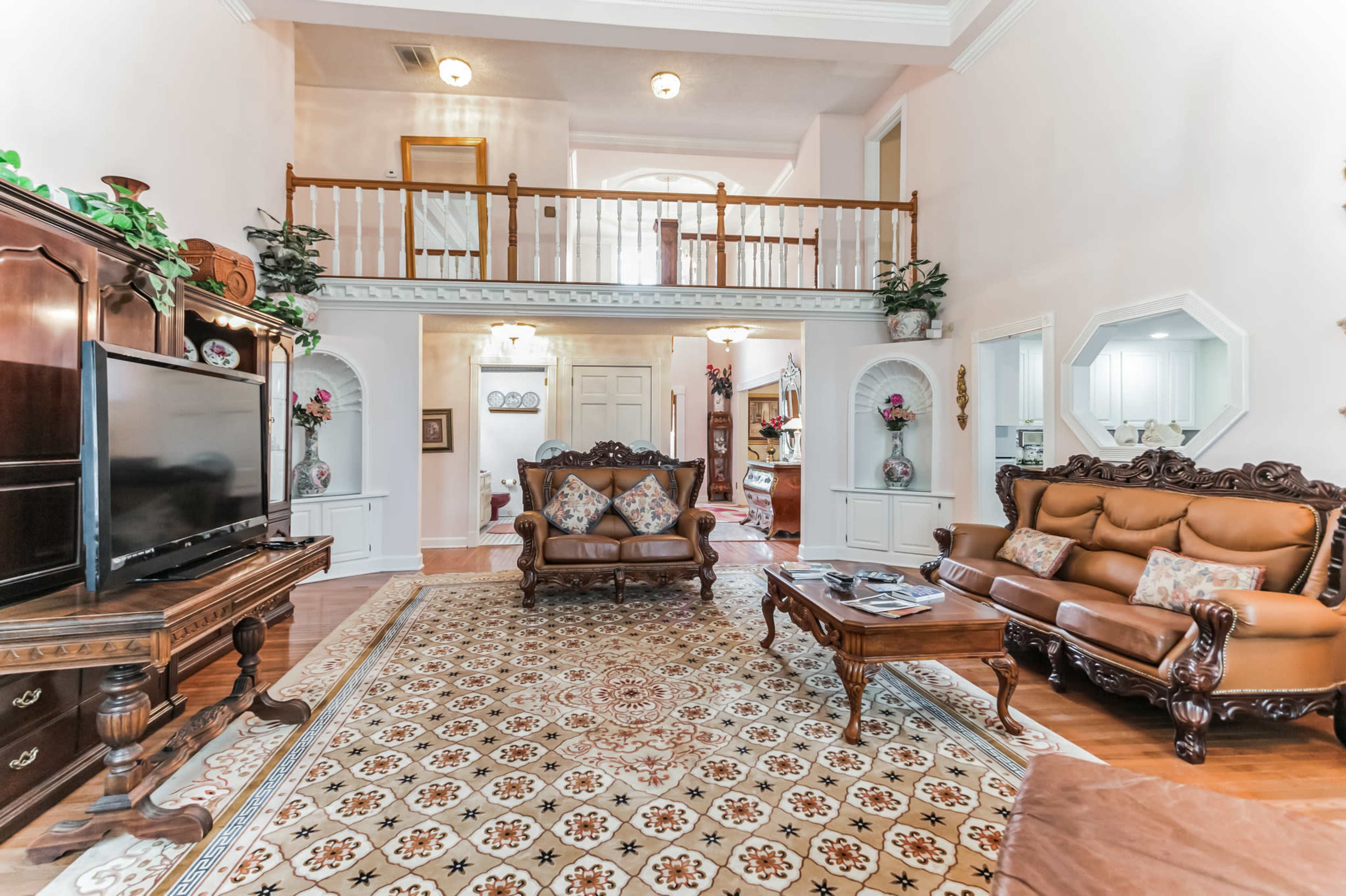A spacious living room with wooden flooring, decorative furniture, a large television, and a balcony overlooking the area.