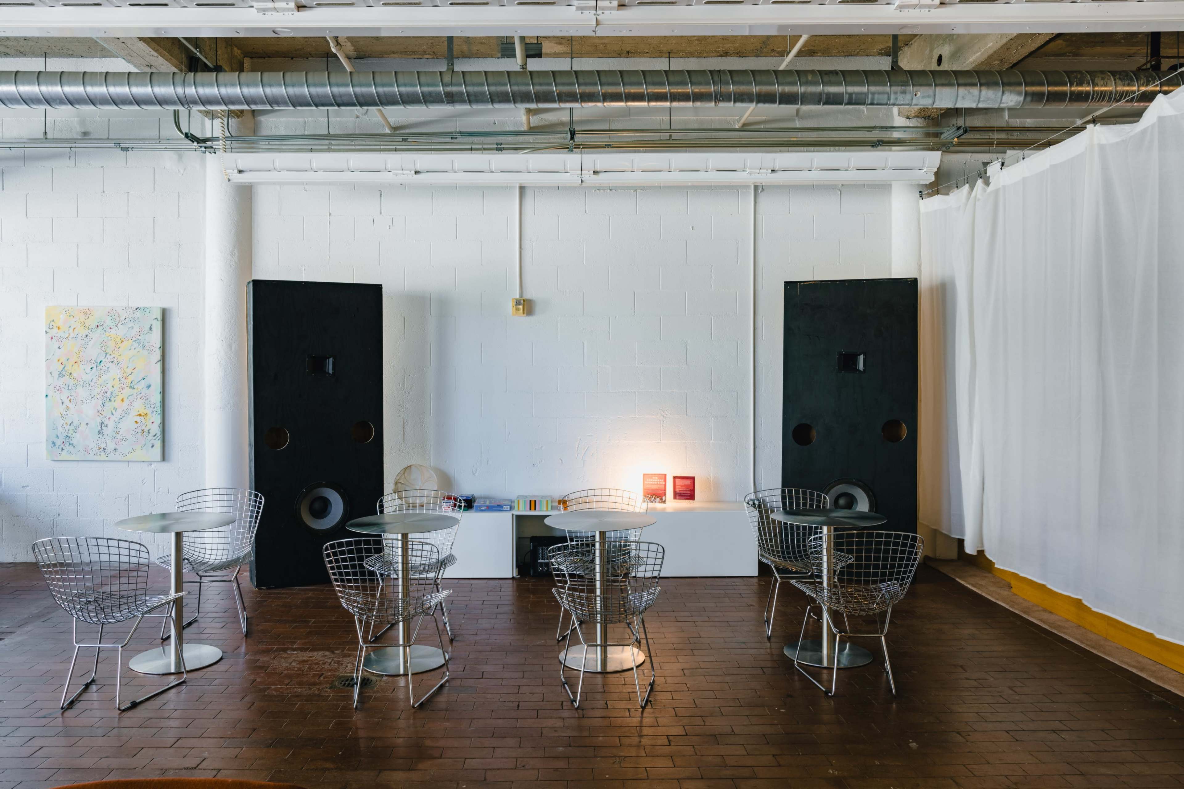 The image shows a minimalist café space with wire chairs, round tables, and large black speakers mounted against a white wall.