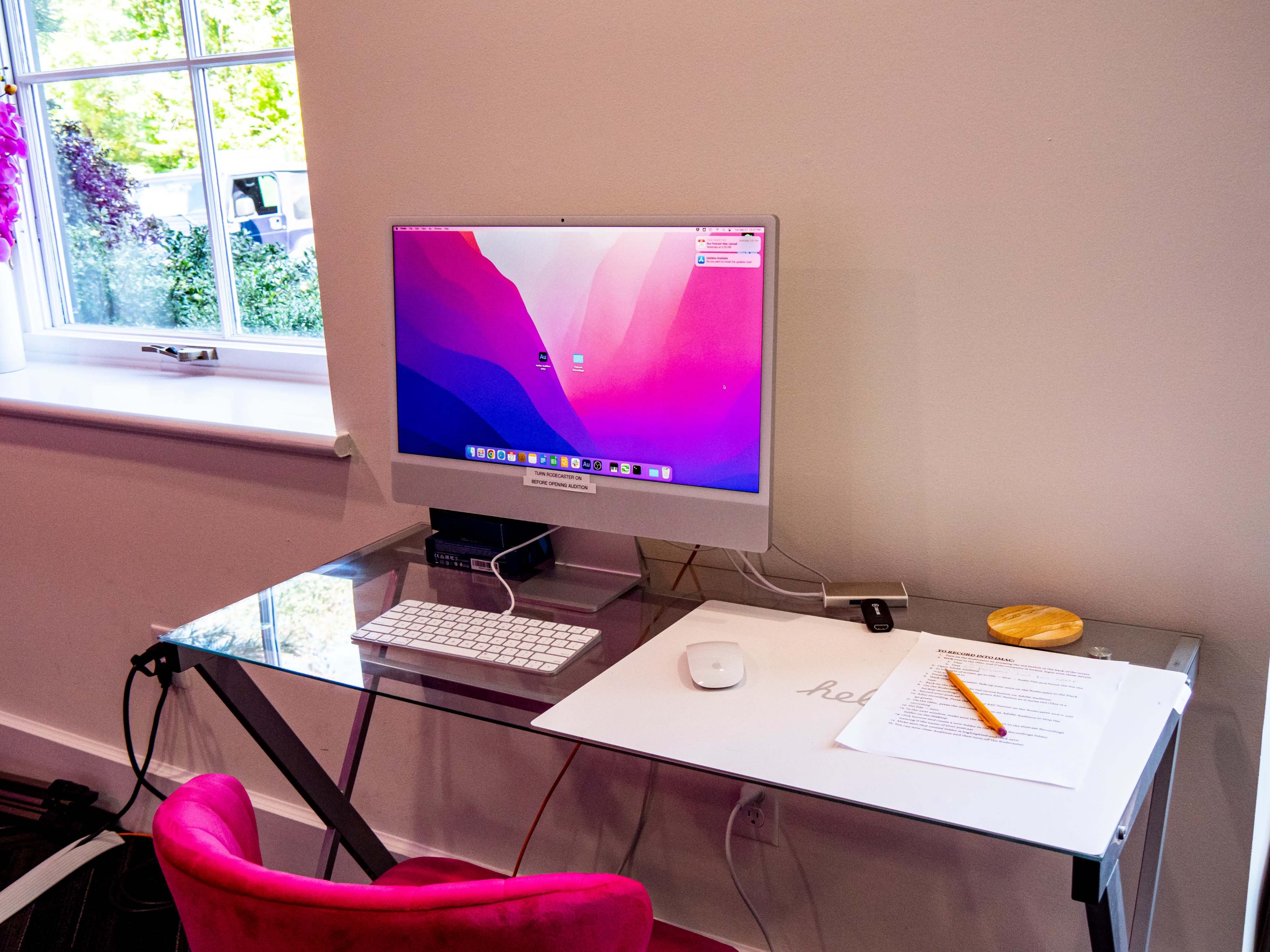 A modern workspace features an Apple computer on a glass desk with a keyboard, mouse, and a document alongside an orange pen.