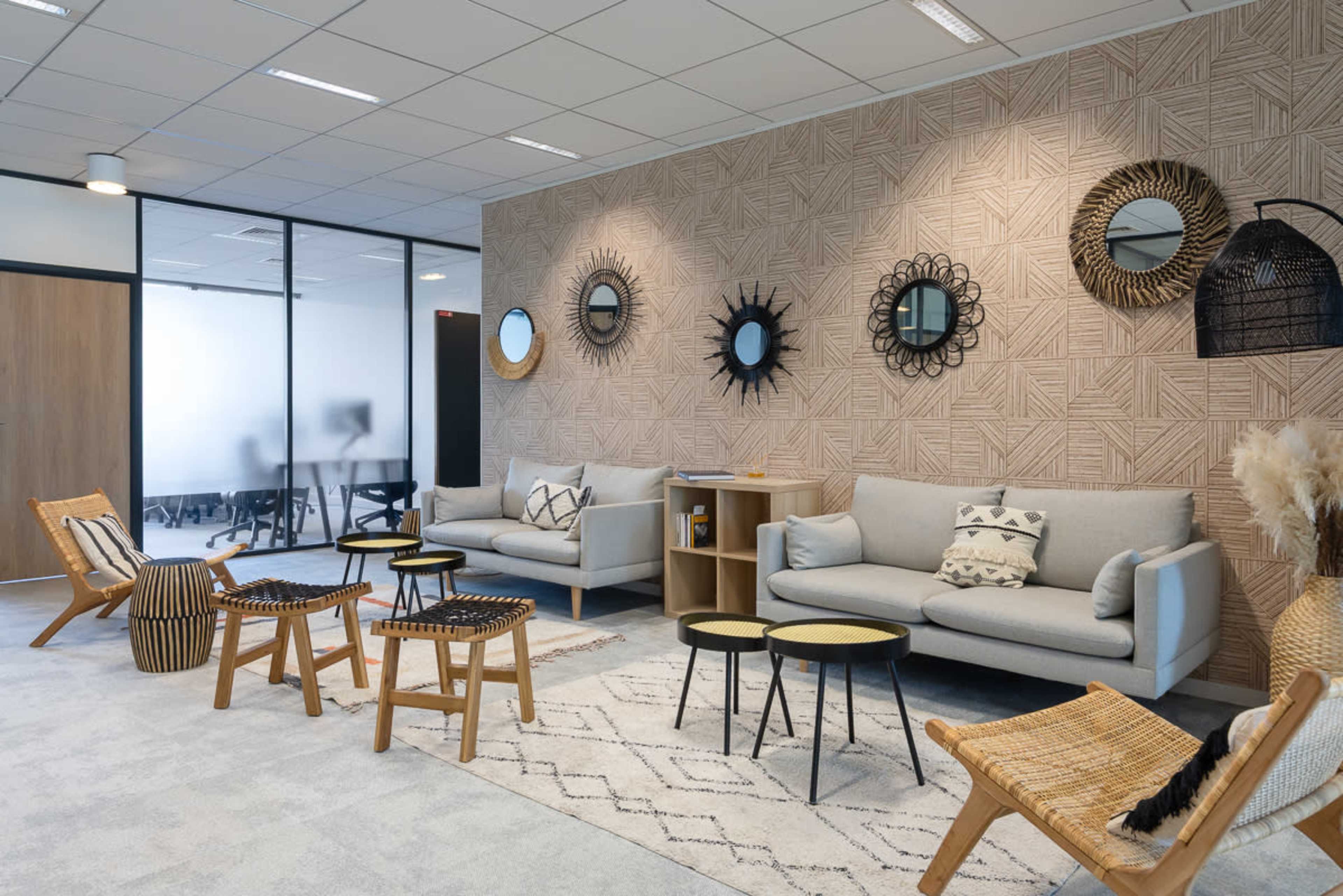 The image shows a modern office lounge area featuring two gray sofas, a patterned wall, decorative mirrors, and a combination of seating options including wicker chairs and small tables.