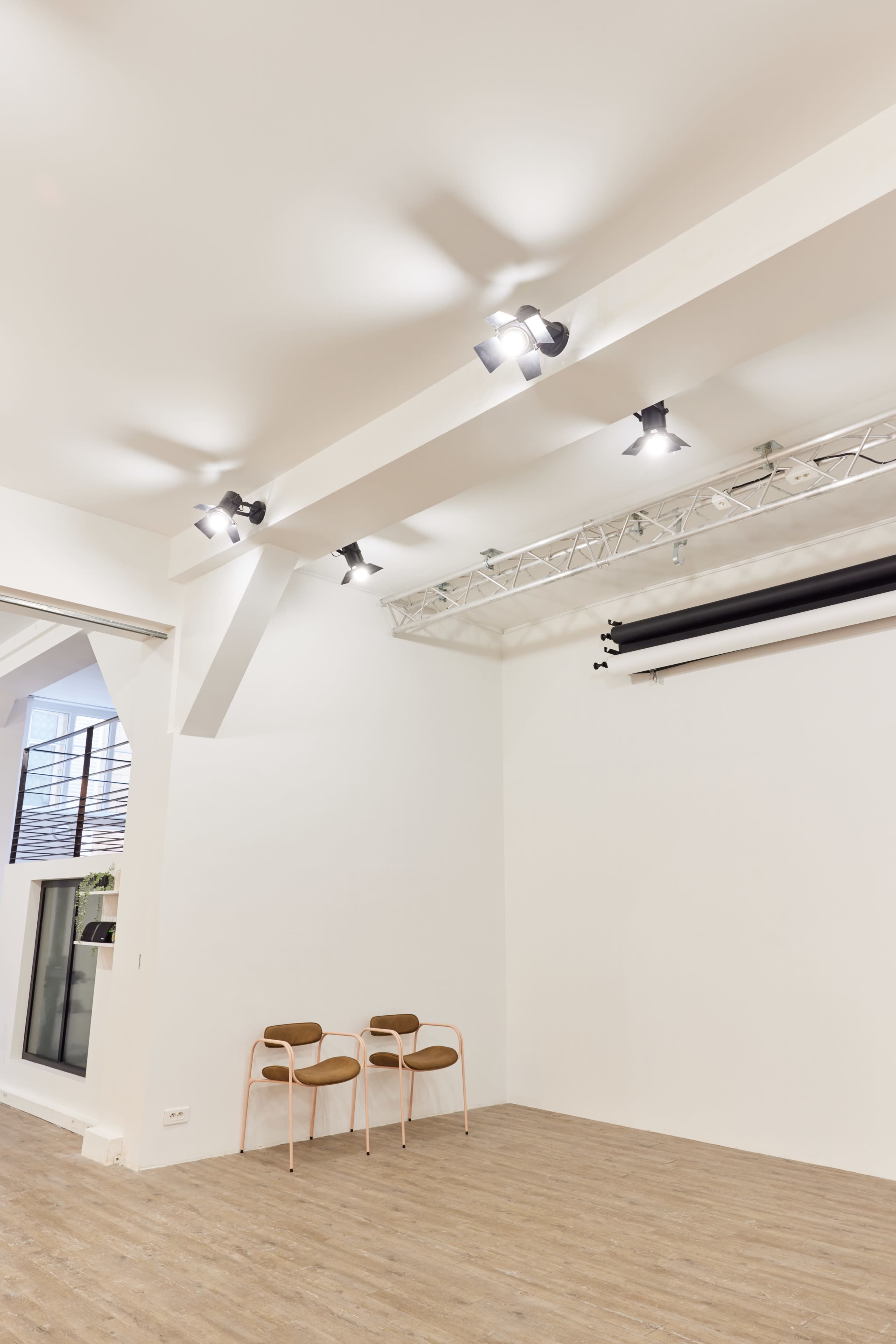 The image shows a minimalist studio space with a white wall, track lighting, and two wooden chairs.