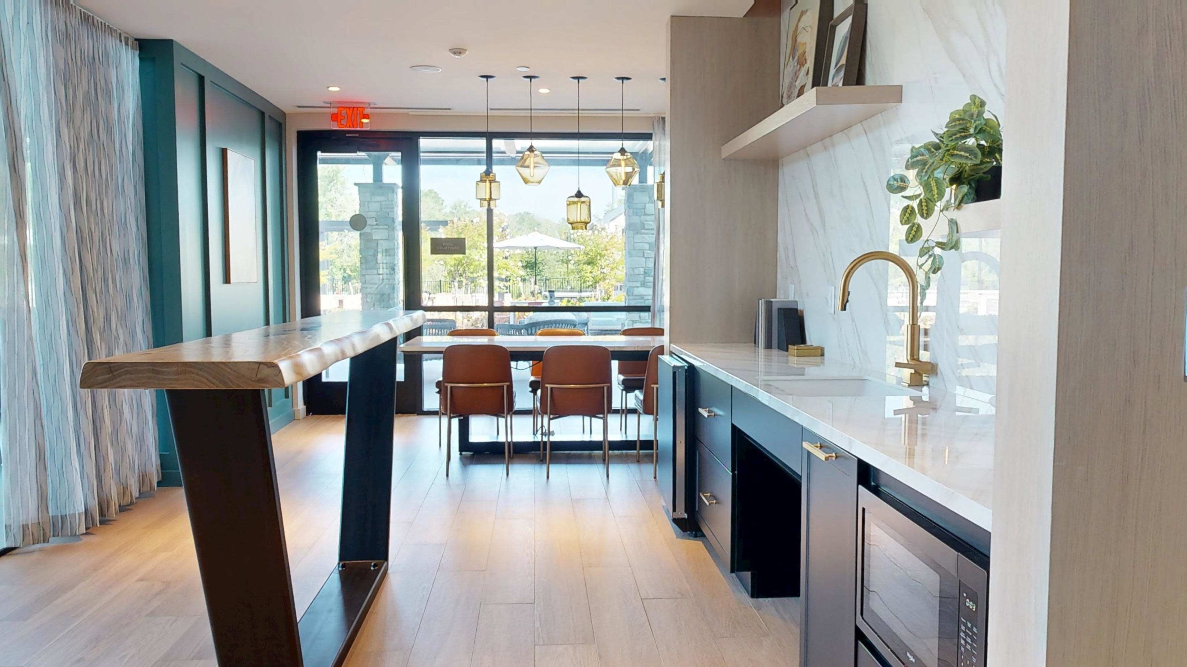 The image shows a modern kitchen area with a sleek countertop, a dining space featuring brown chairs, and large windows allowing natural light to fill the room.
