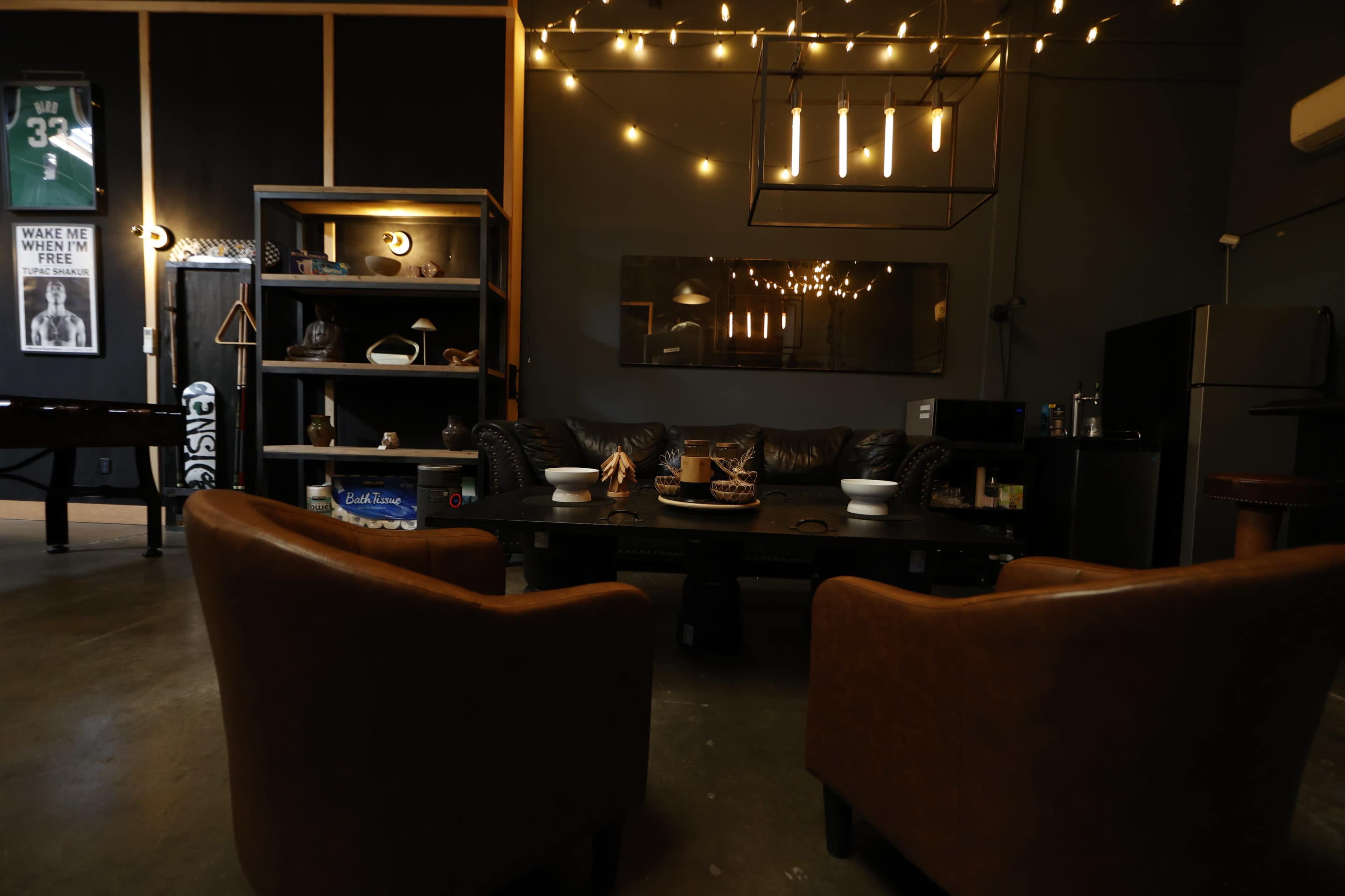 The image features a stylish room with a dark color scheme, a leather couch, two brown chairs, decorative string lights, and various items on display.