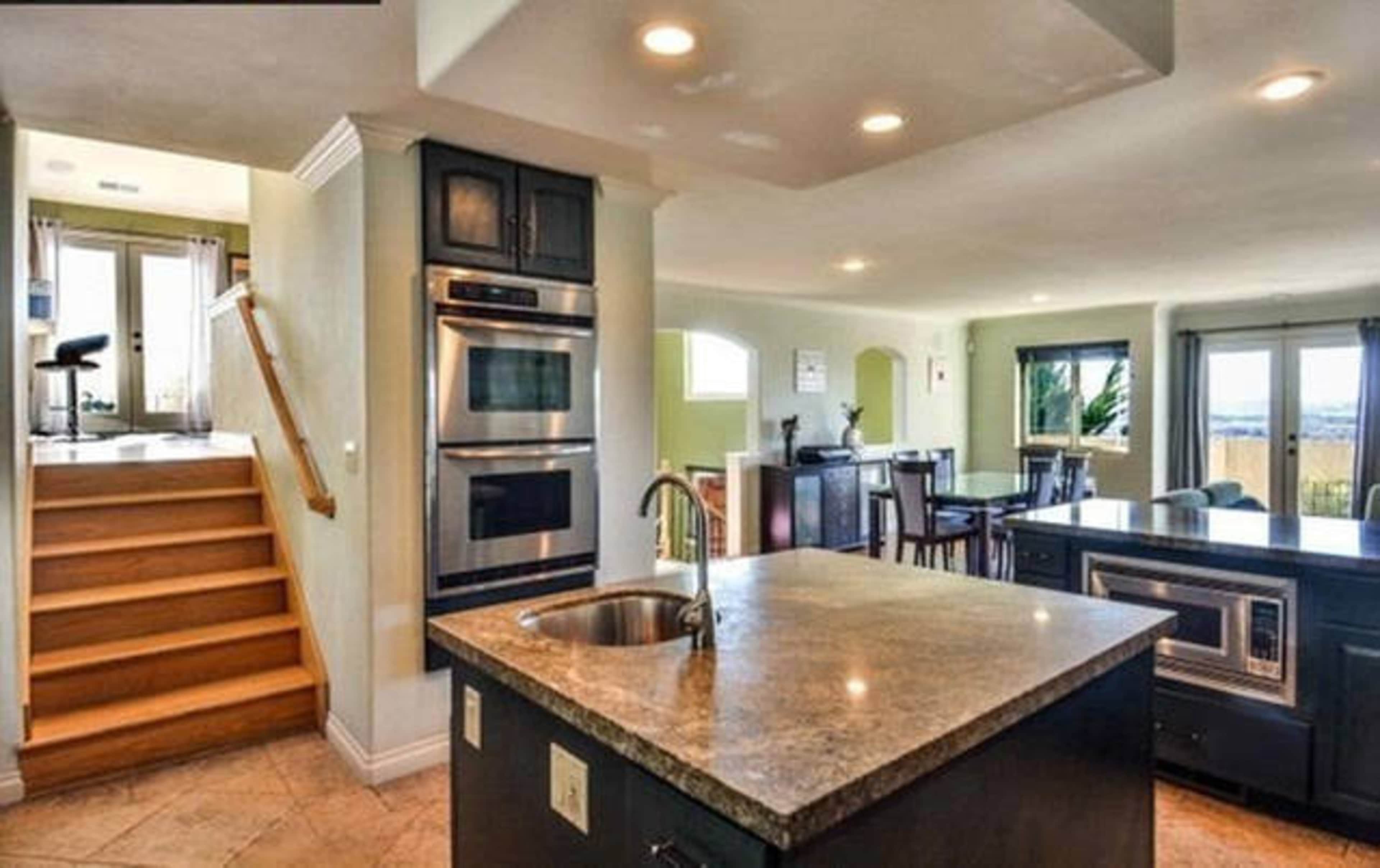 The image shows a modern kitchen with an island, stainless steel appliances, and wooden stairs leading to an upper level.