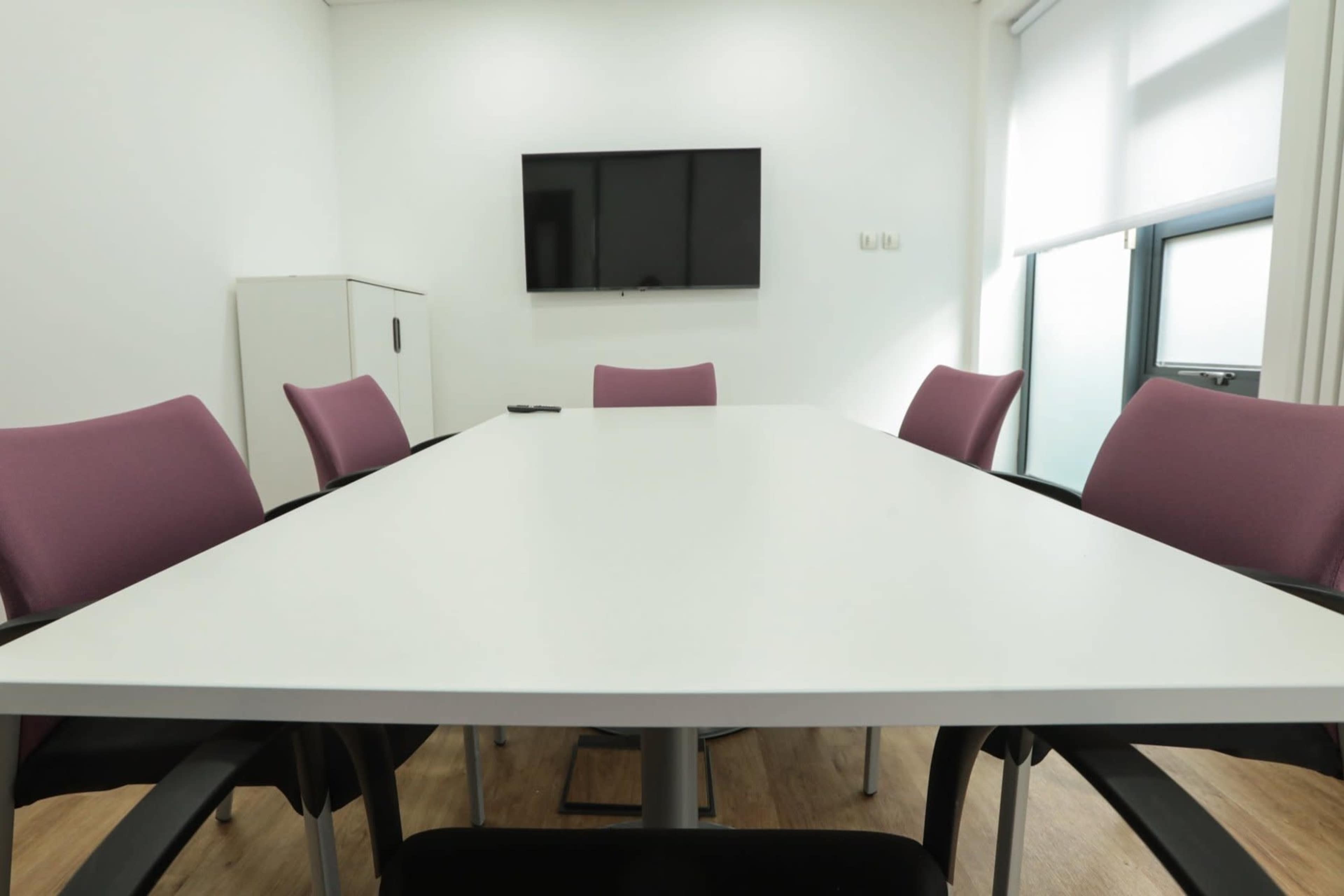 A modern conference room features a large white table surrounded by purple chairs, with a wall-mounted television in the background.