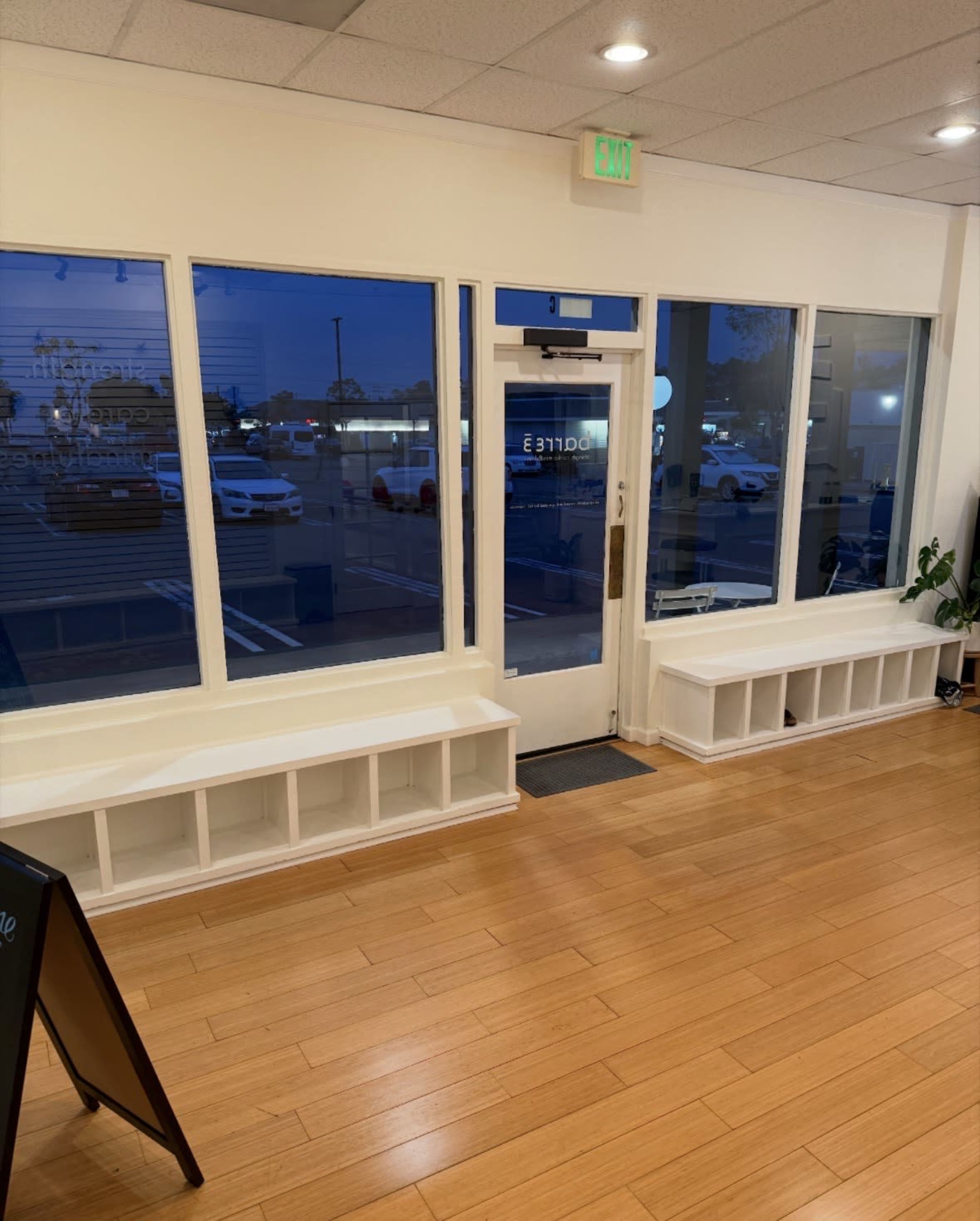 The image shows the interior of a store featuring large windows, a front door, and two built-in benches along the walls, with a view of parked cars outside during twilight.