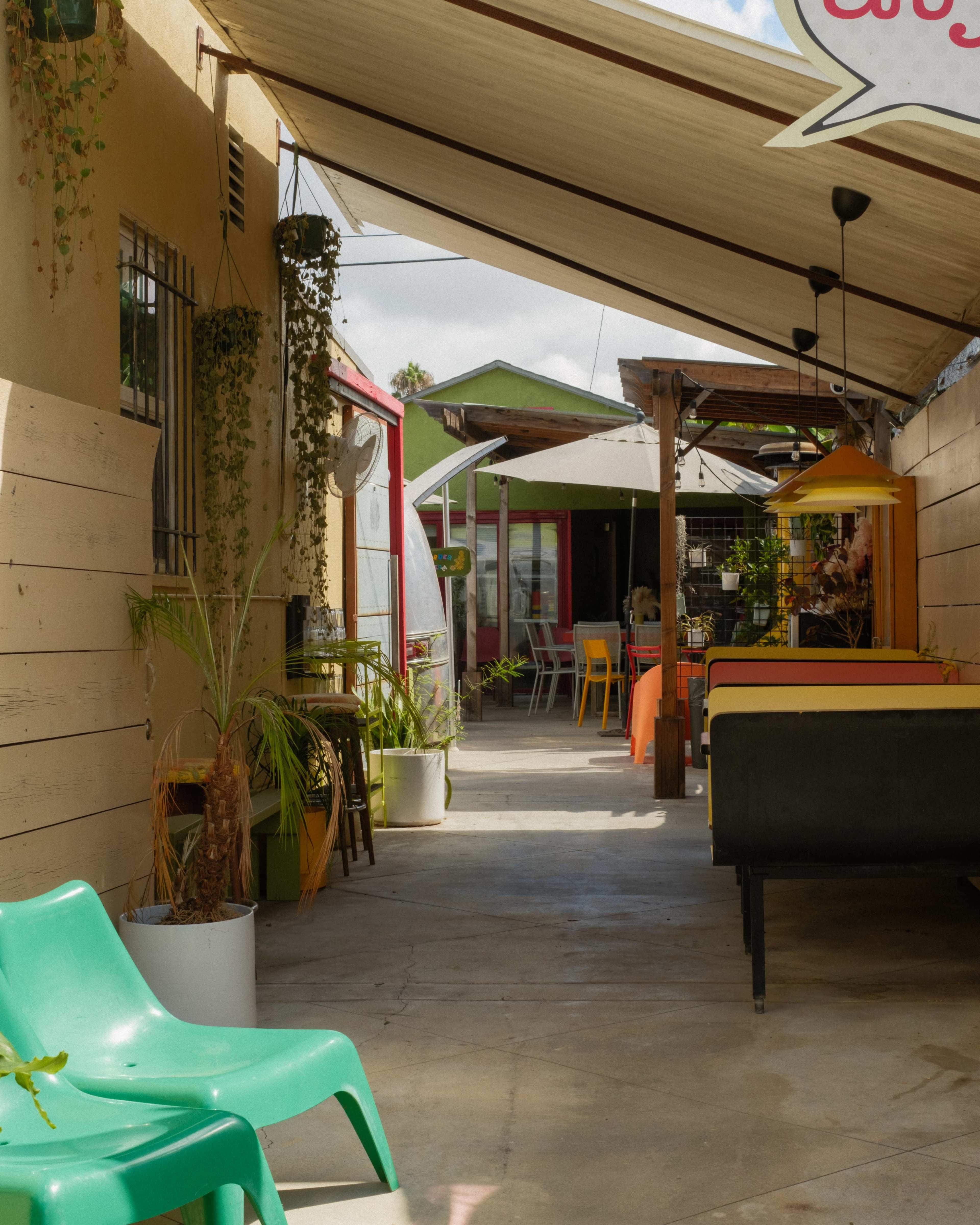 The image shows a narrow outdoor passage with colorful chairs and tables, lined with plants and shaded by a canopy.