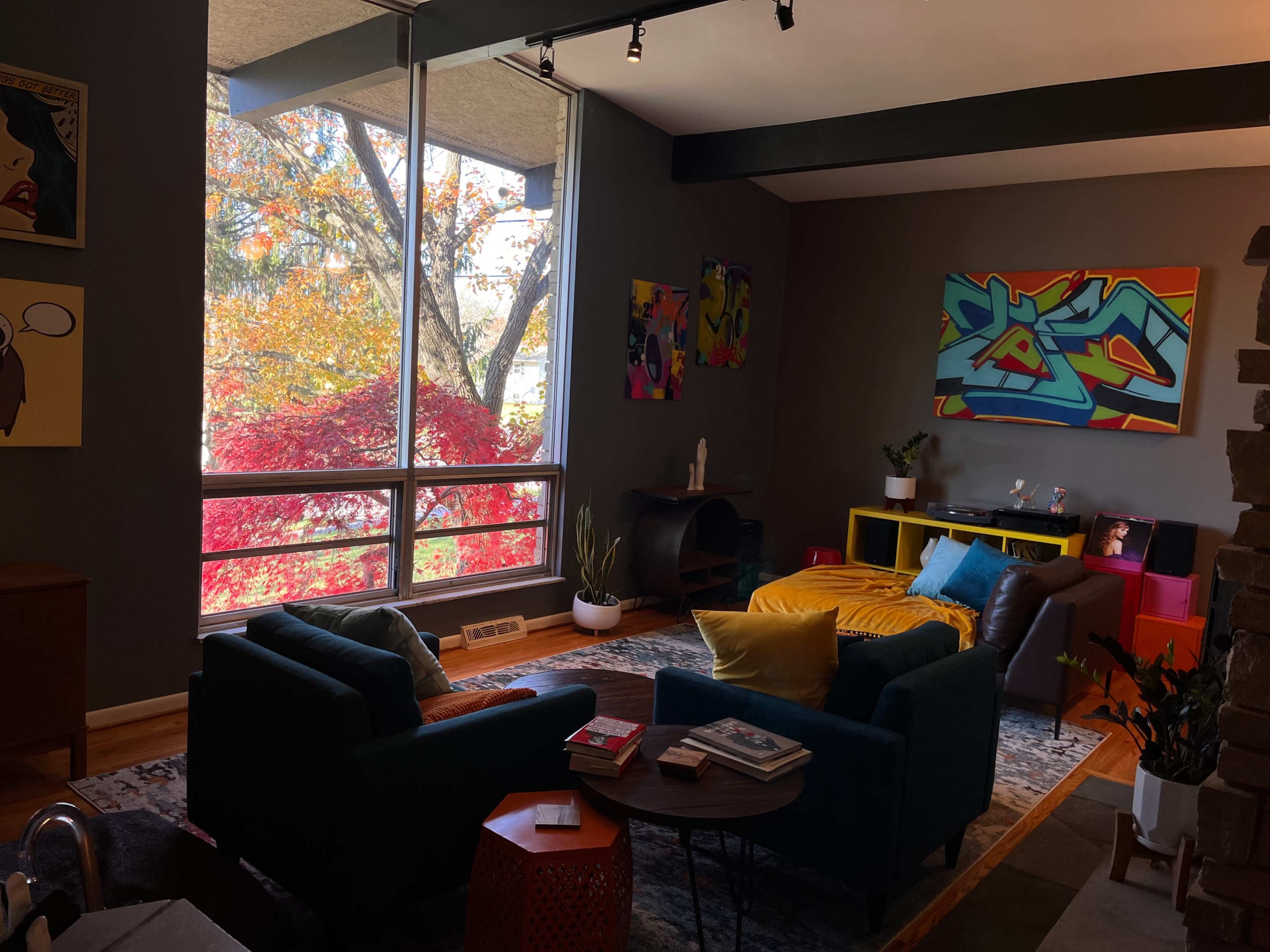 A modern living room with two teal sofas, a colorful rug, a coffee table, and large windows displaying autumn foliage outside.