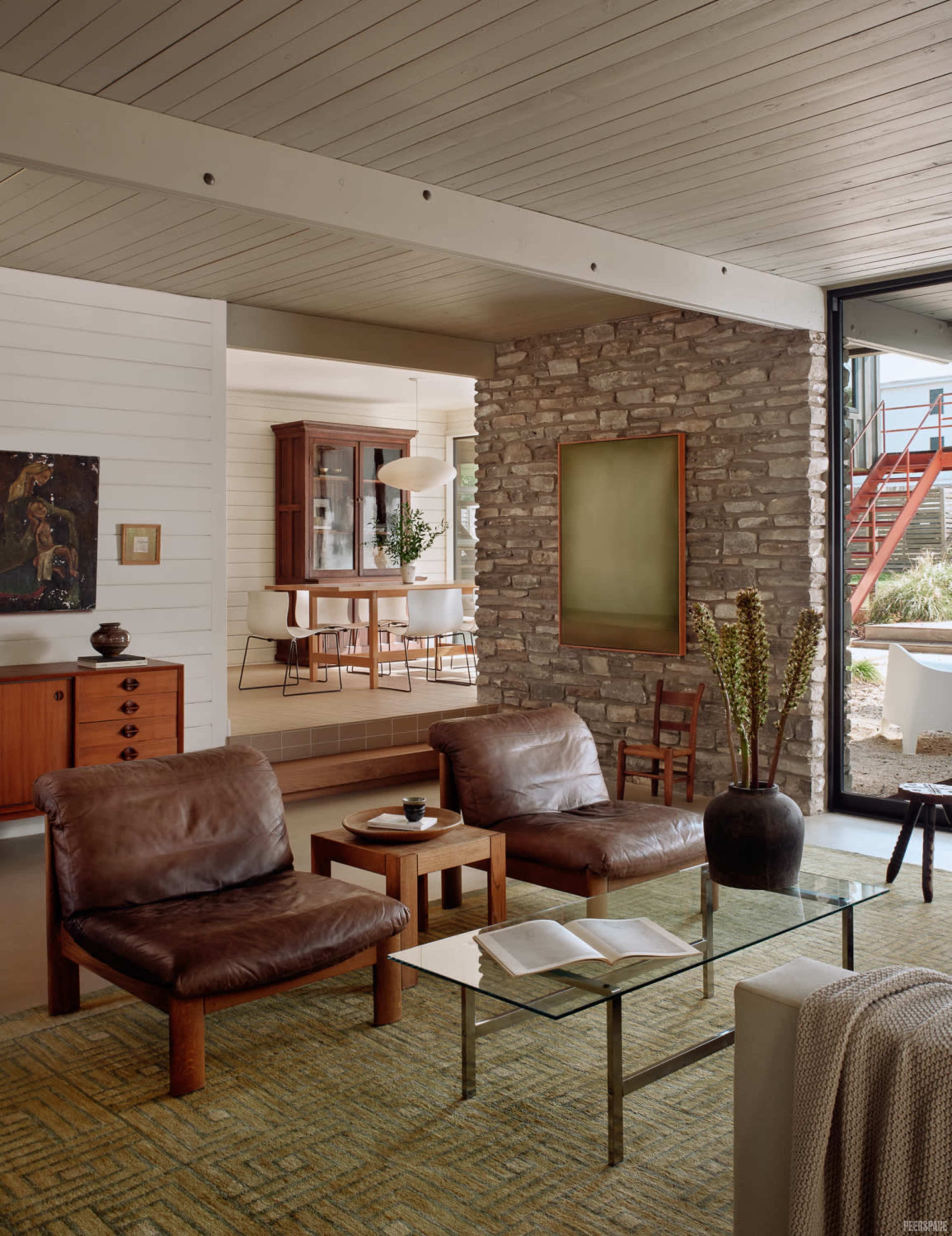 The image shows a modern living room with brown leather chairs, a glass coffee table, and a stone accent wall, leading to a dining area in the background.