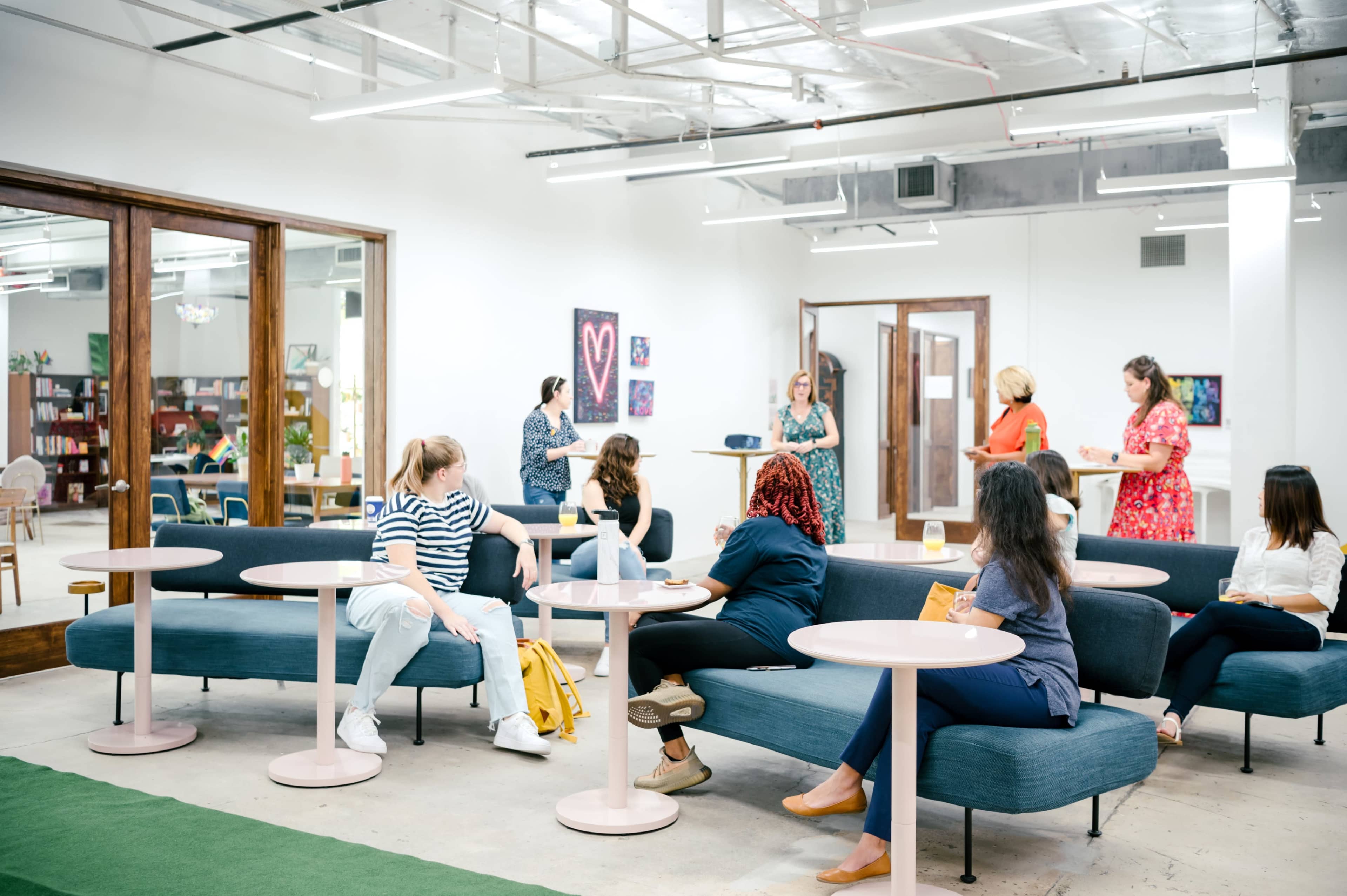 A group of women are engaged in conversation and activities in a modern, open workspace furnished with casual seating and artwork on the walls.