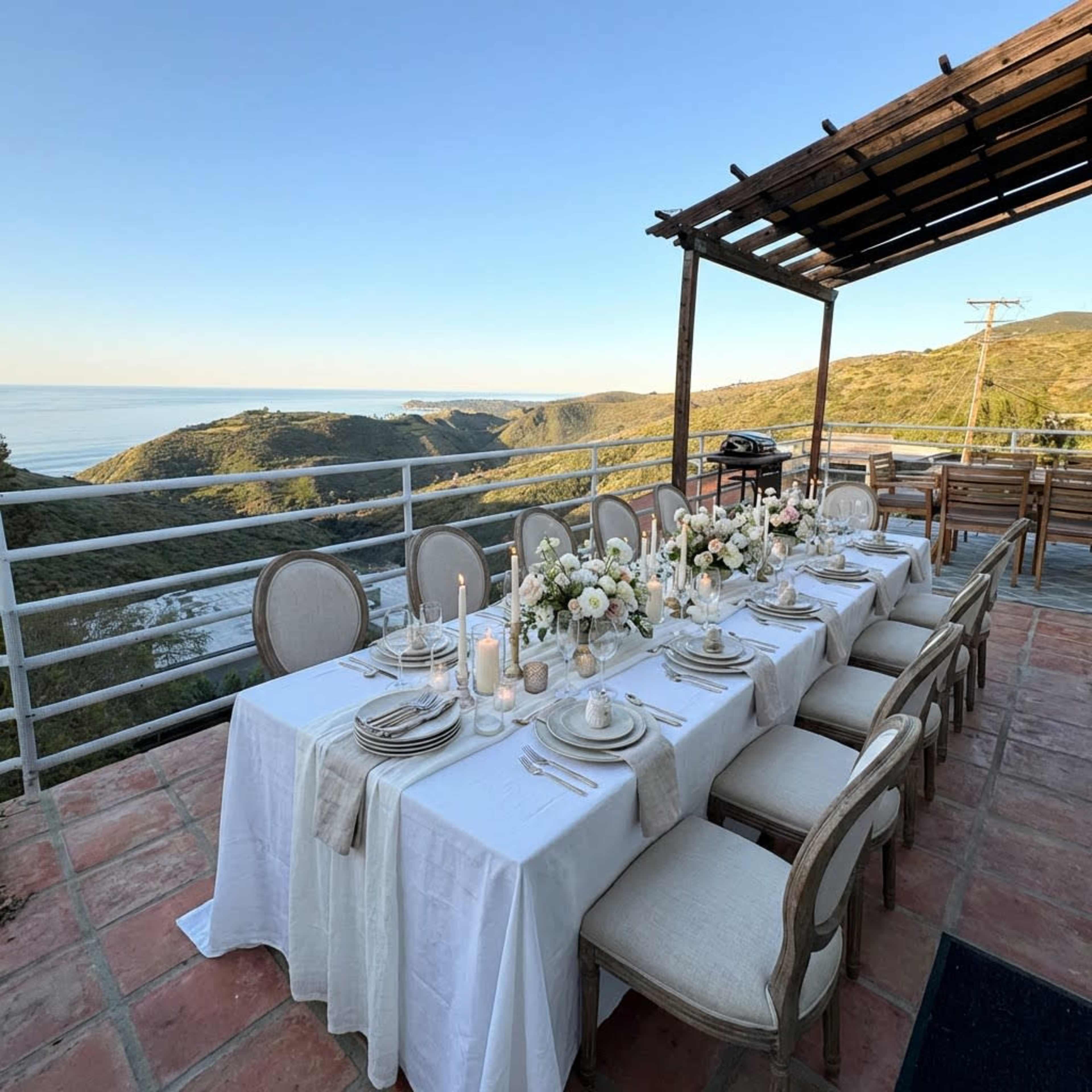 A long dining table is set for an outdoor meal overlooking a scenic coastline and hills.