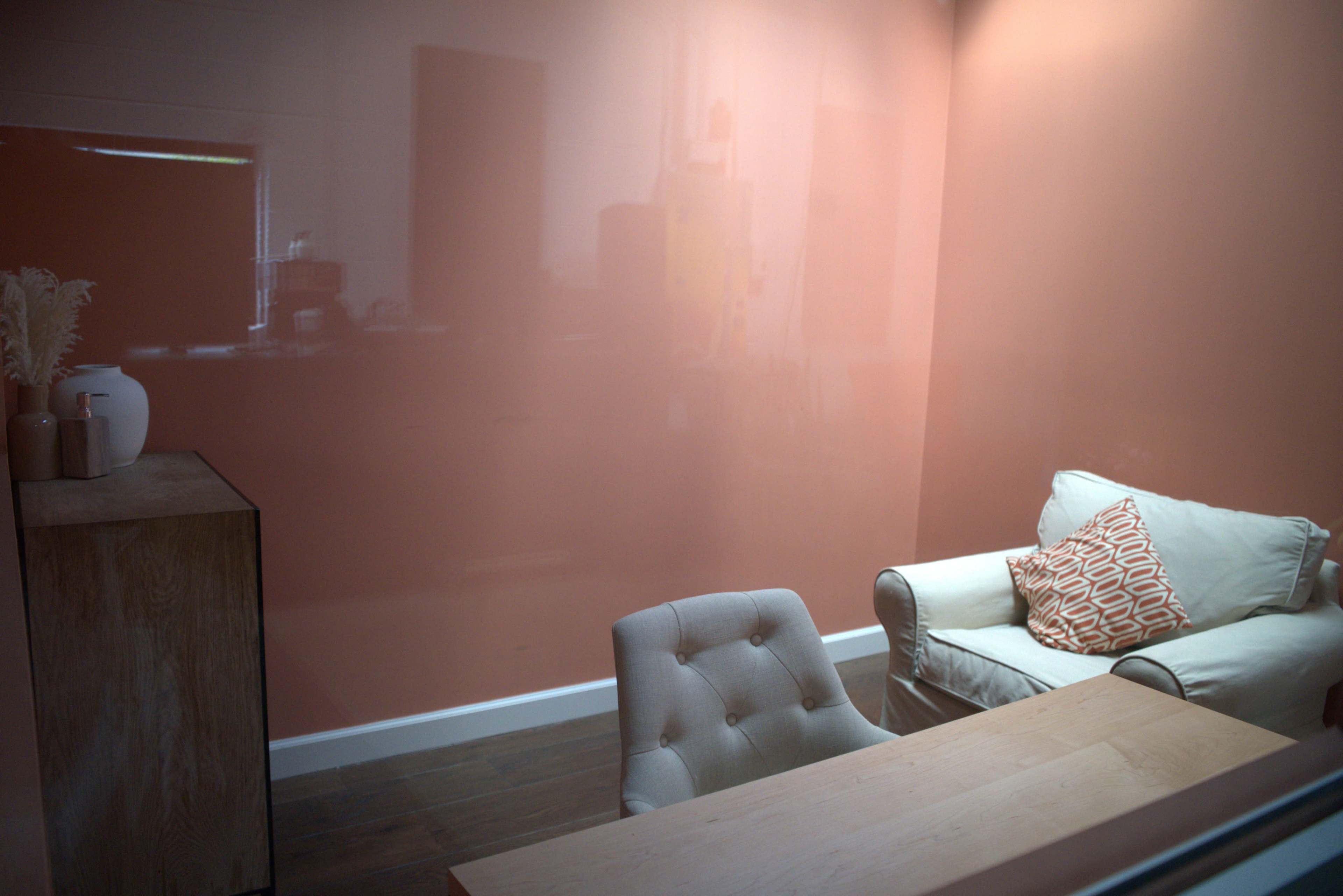 The image shows a small room with a peach-colored wall, featuring a beige armchair, a wooden desk, and a decorative plant on a side table.