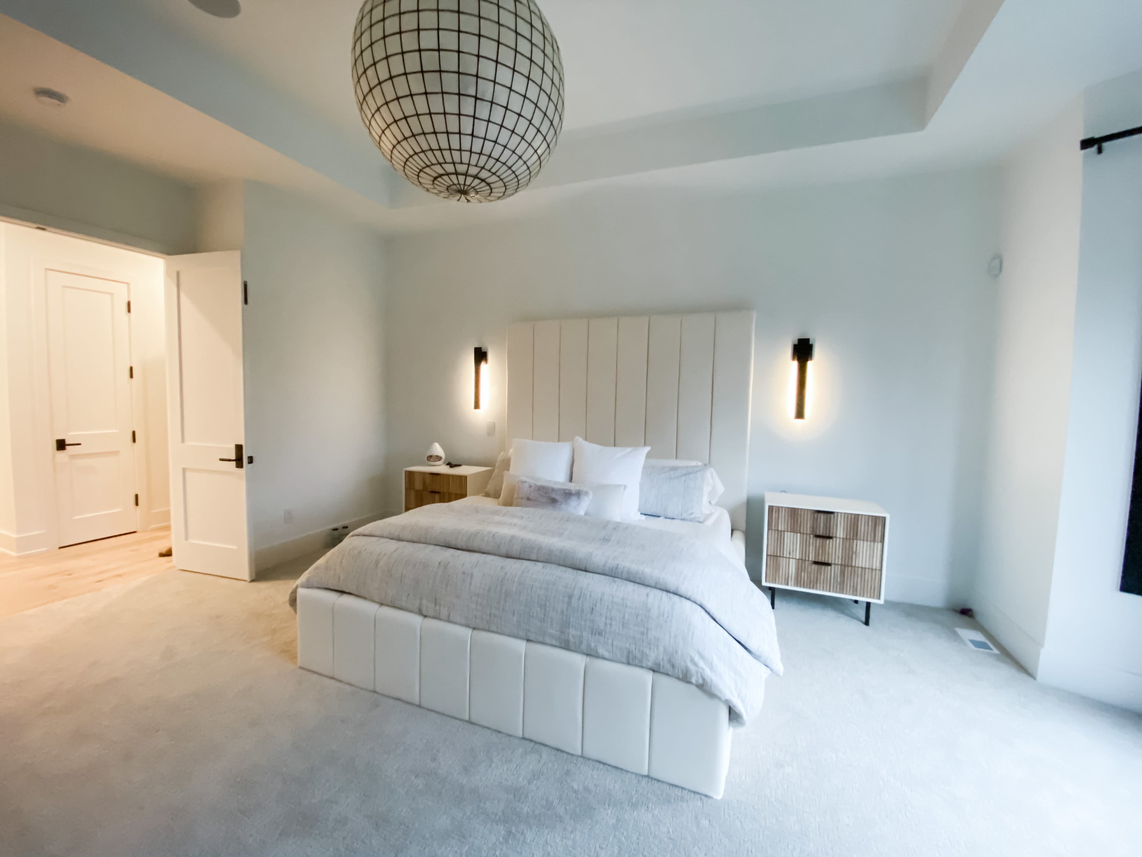 The image shows a modern bedroom featuring a large bed with a light gray blanket, two wall-mounted lights, a round pendant lamp, and a wooden nightstand.