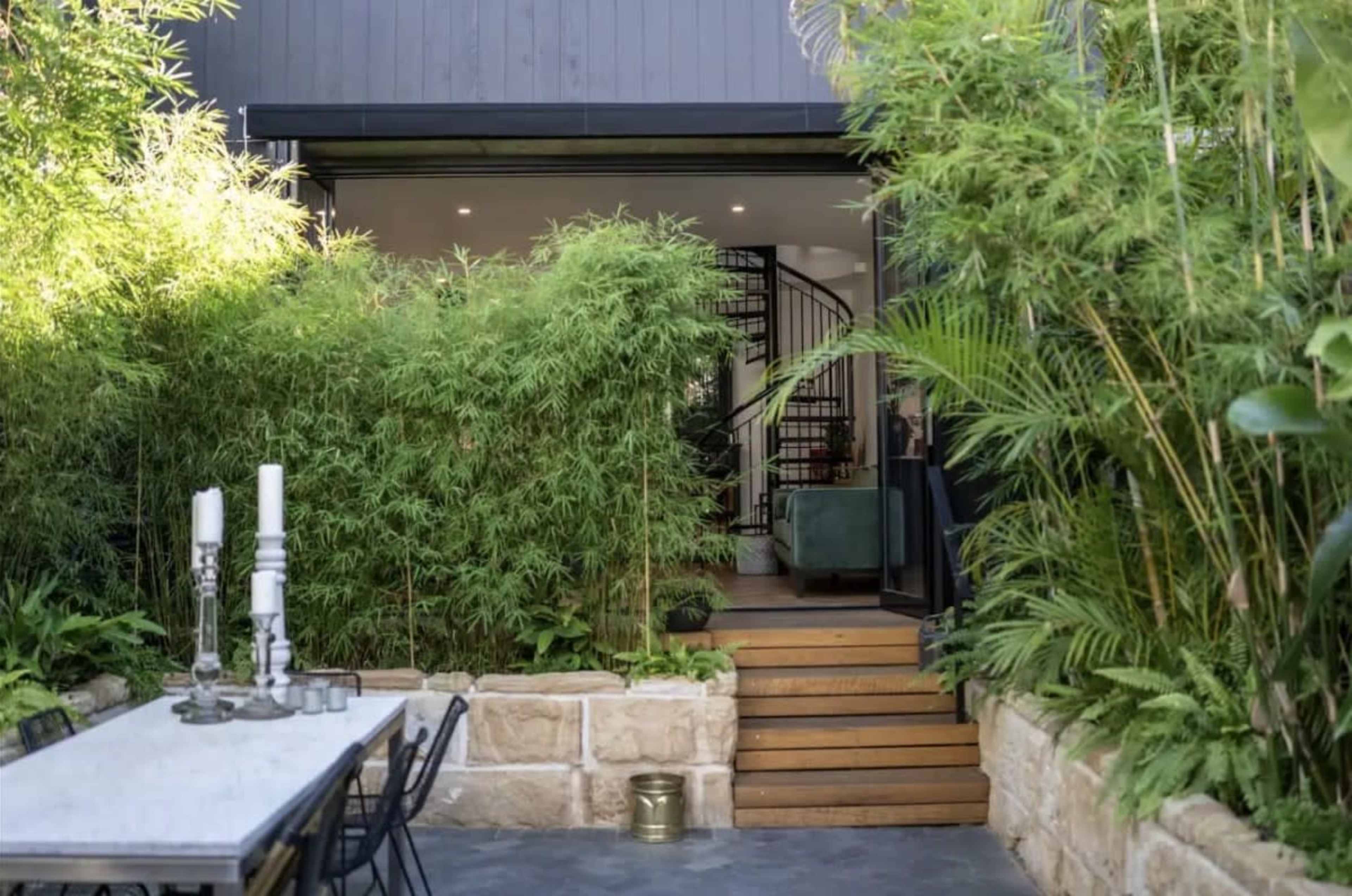 A set of wooden stairs leads from a stone patio through a verdant garden towards a modern entrance.