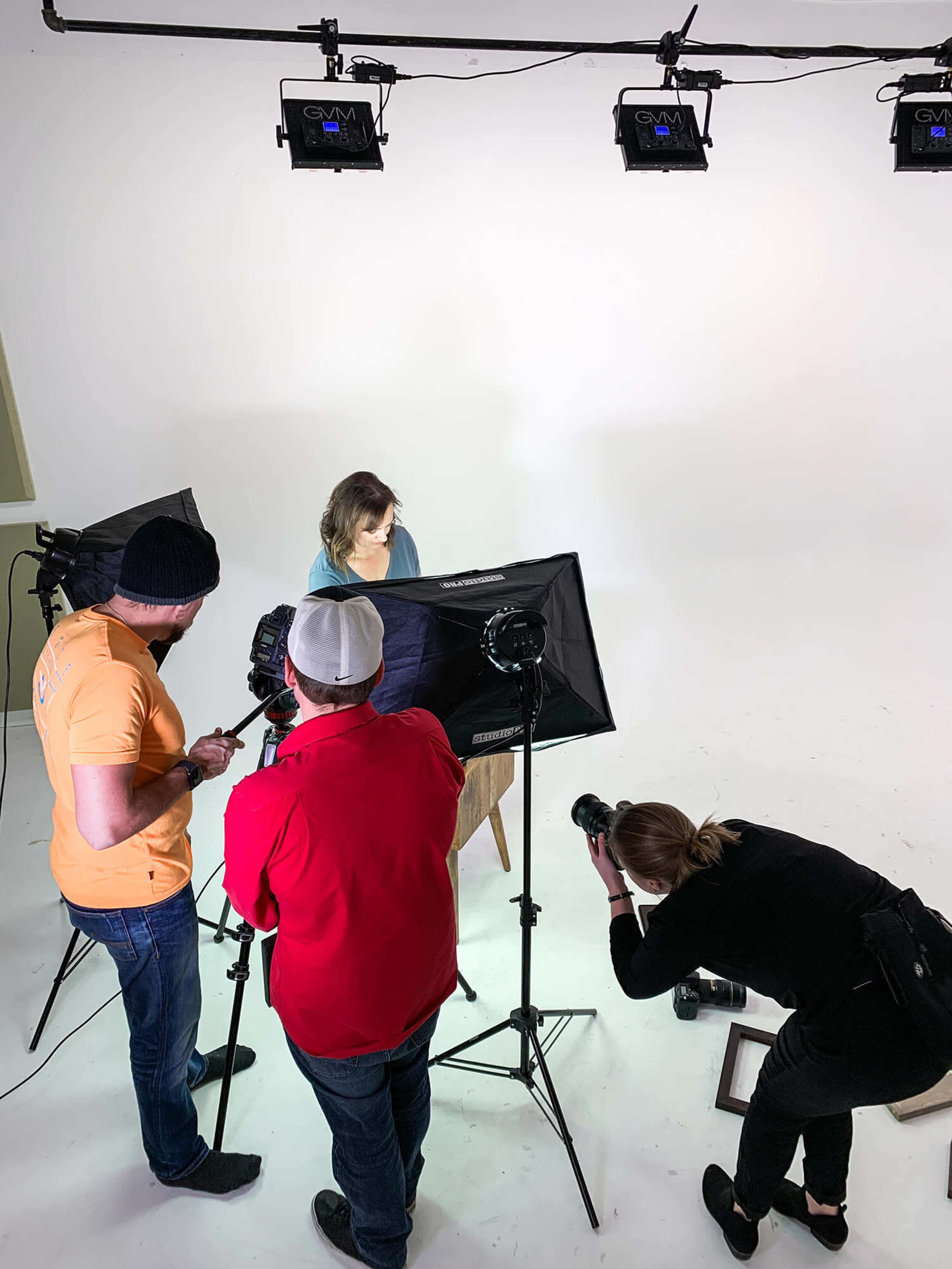 A small film crew is setting up equipment around a woman seated on a chair in a brightly lit studio.