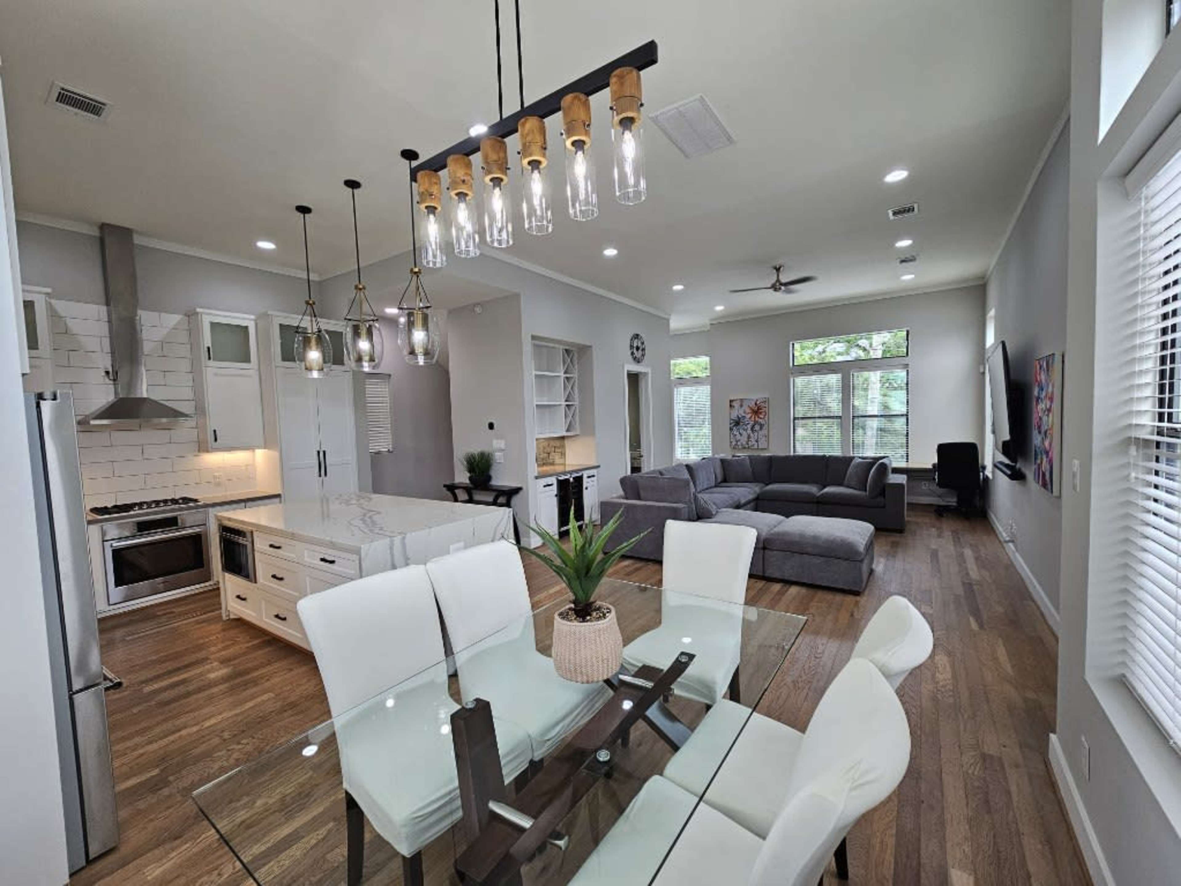 A modern open-concept living space featuring a dining area with a glass table, white chairs, a kitchen with stainless steel appliances, and a living area with a gray sectional sofa.