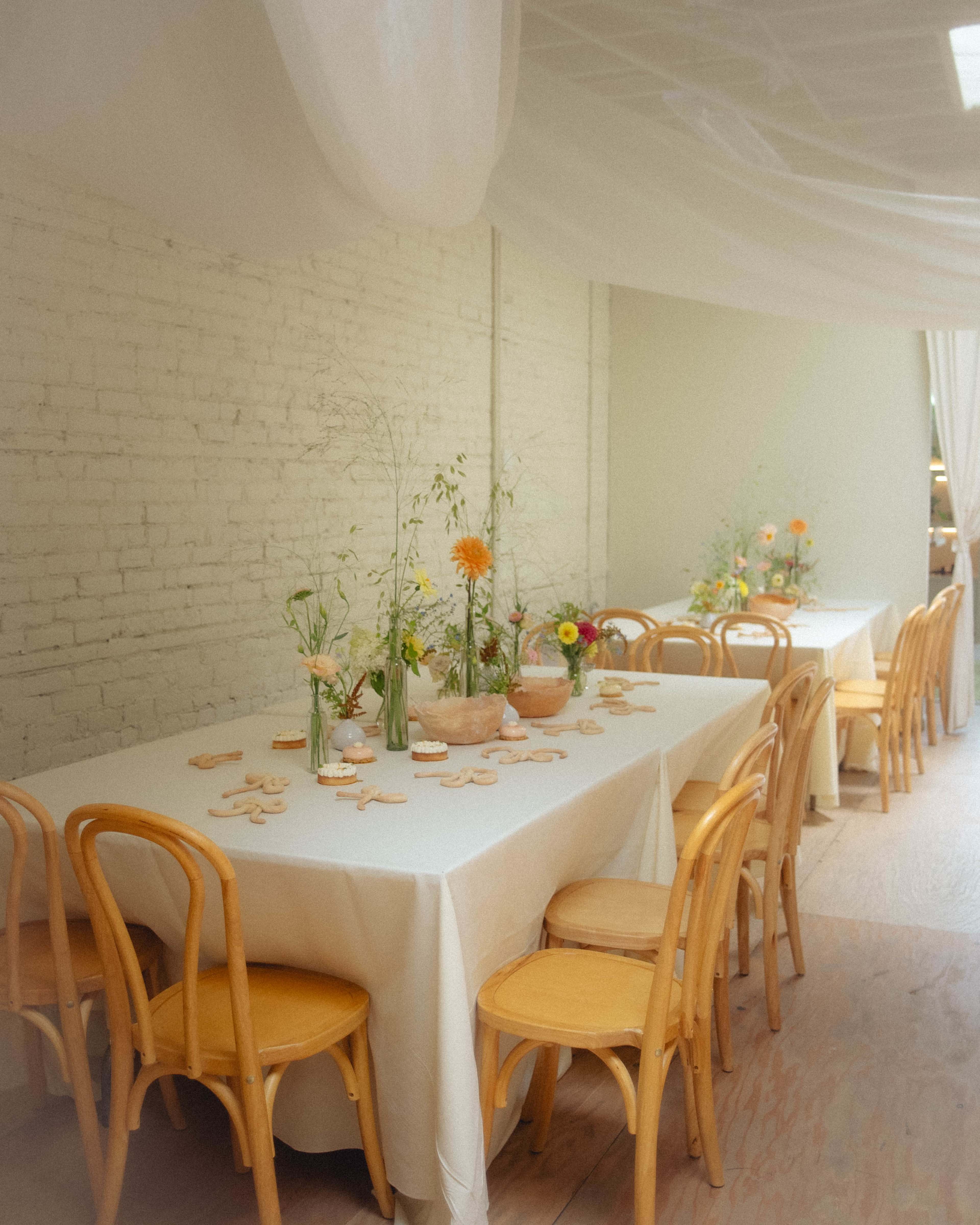 A bright room features a long dining table set with simple floral arrangements and wooden chairs.