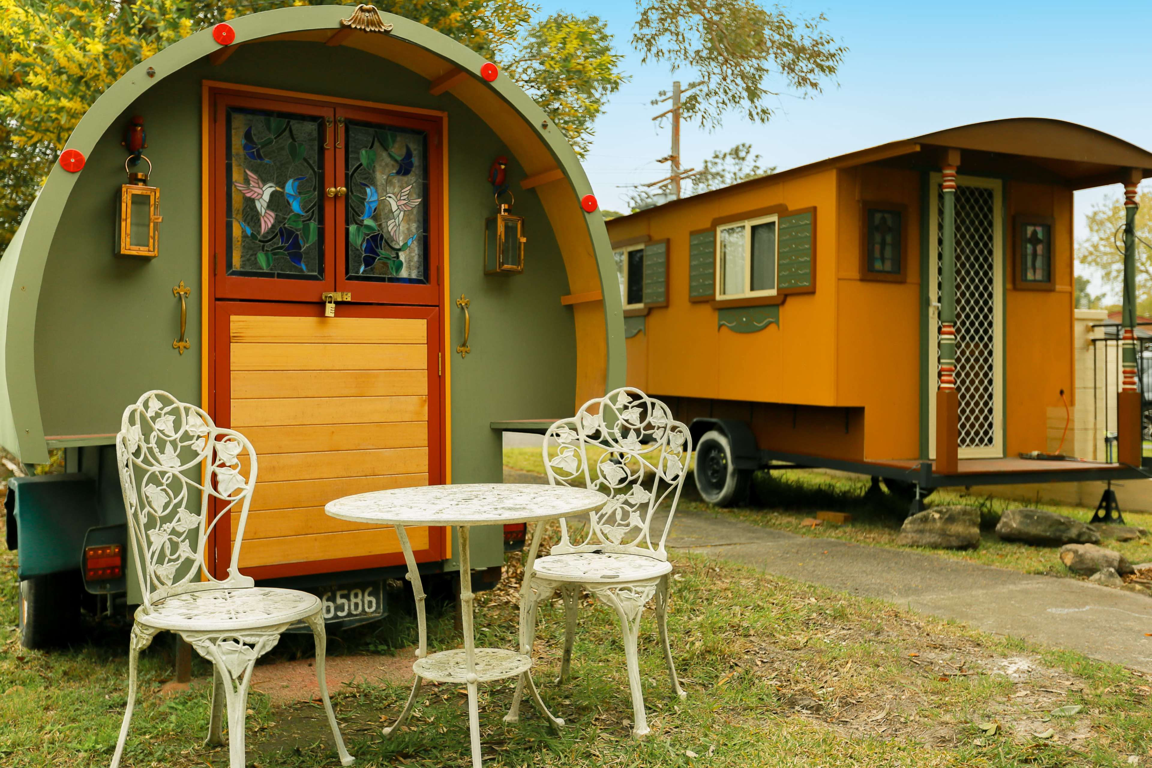 A colorful vintage trailer is parked next to a small cabin, with a white table and chairs set up in front of the trailer.