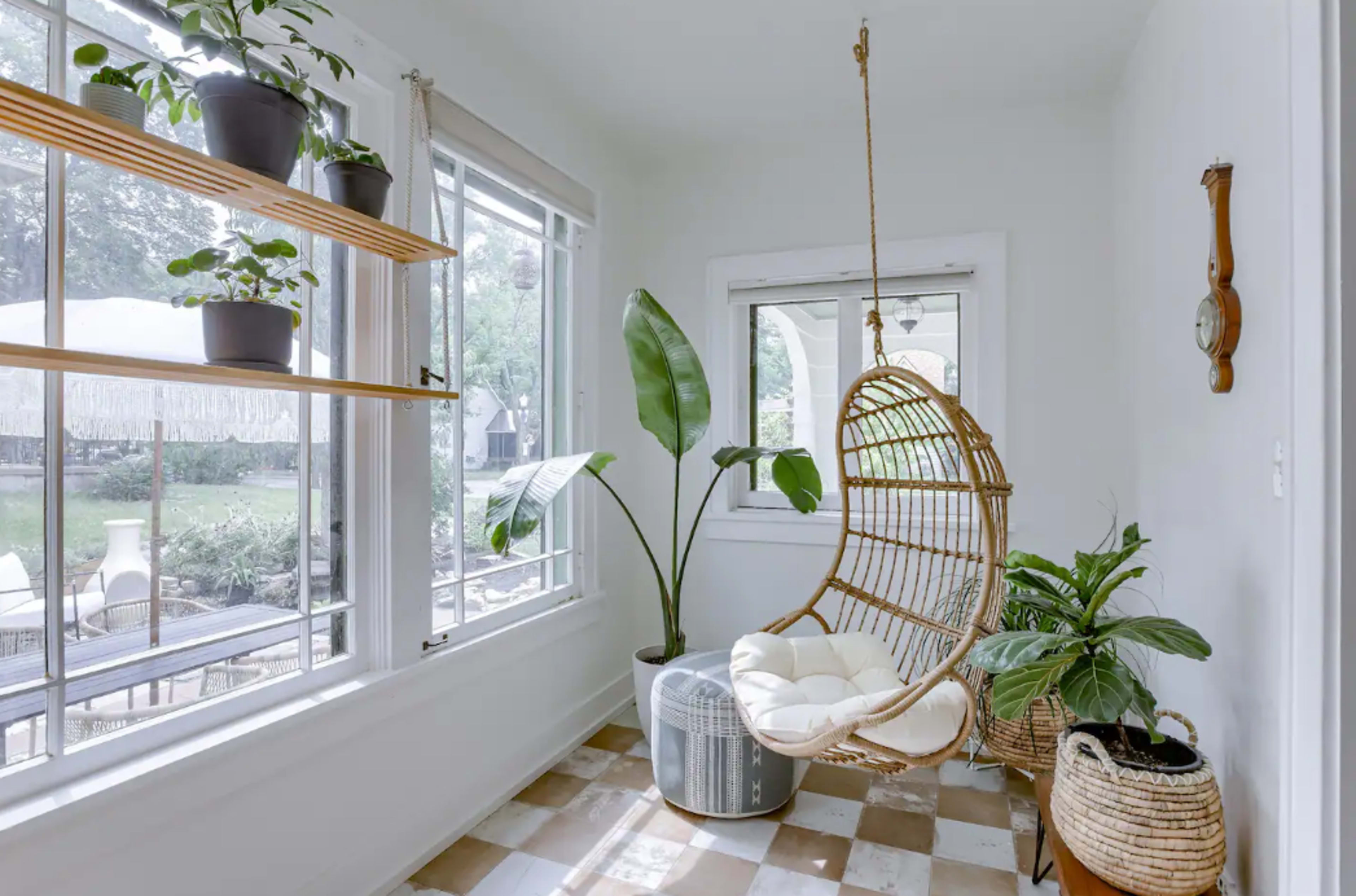 A sunlit corner of a room featuring a hanging rattan chair, indoor plants, and large windows overlooking a garden.