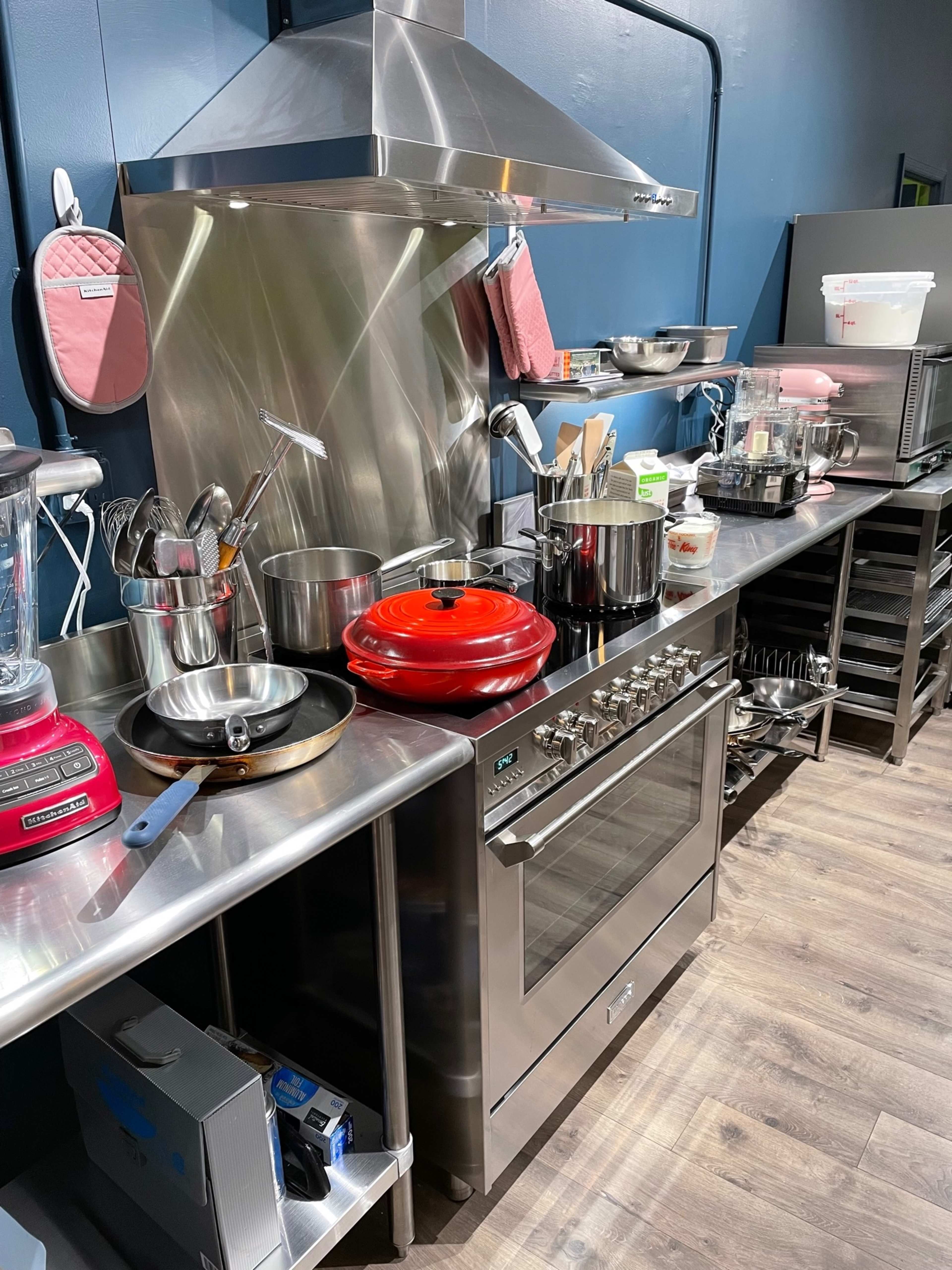 A stainless steel kitchen countertop with various pots, utensils, and appliances, including a red Dutch oven and a professional oven.