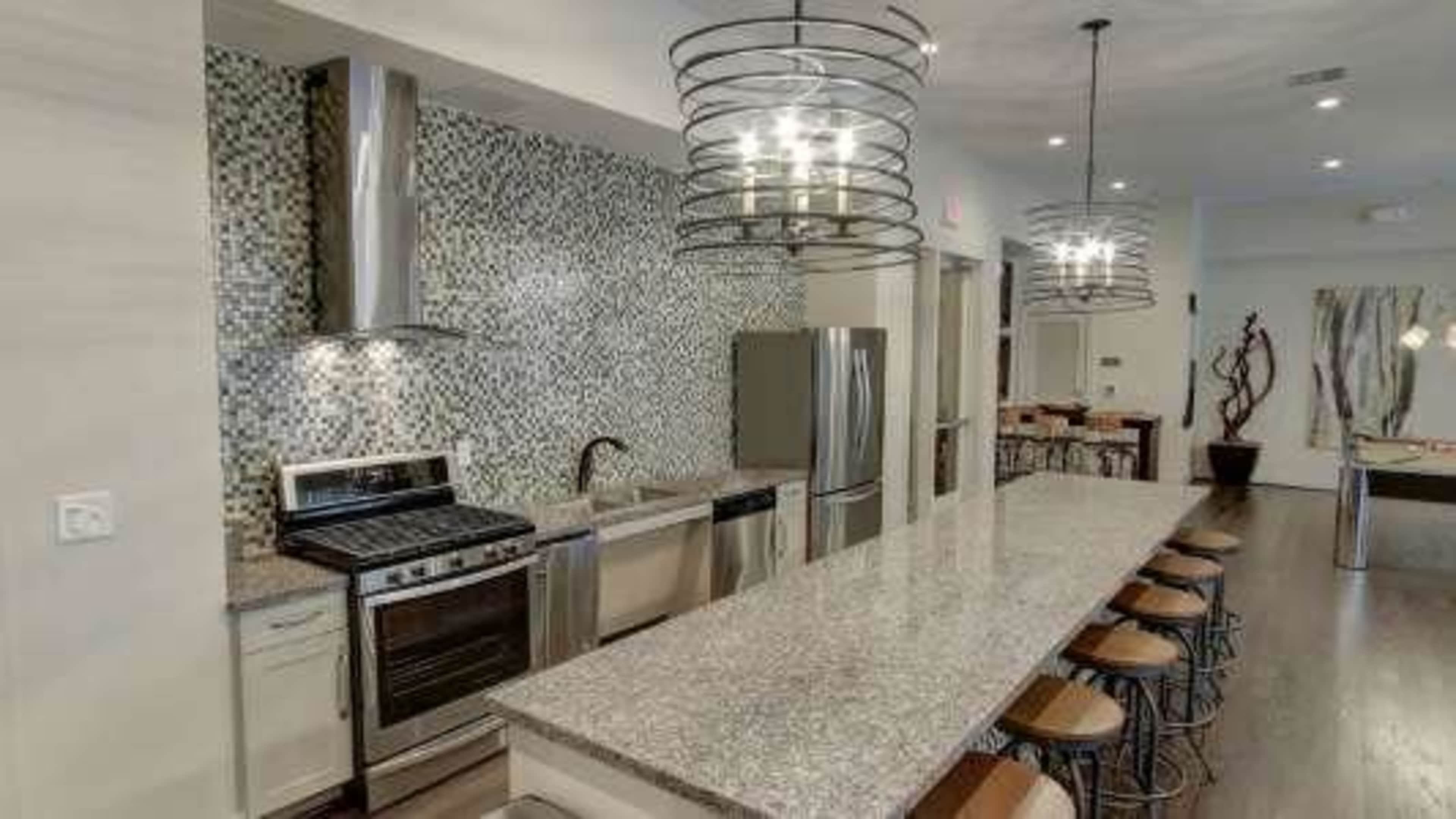 The image shows a modern kitchen with stainless steel appliances, a large granite island, and contemporary light fixtures overhead.