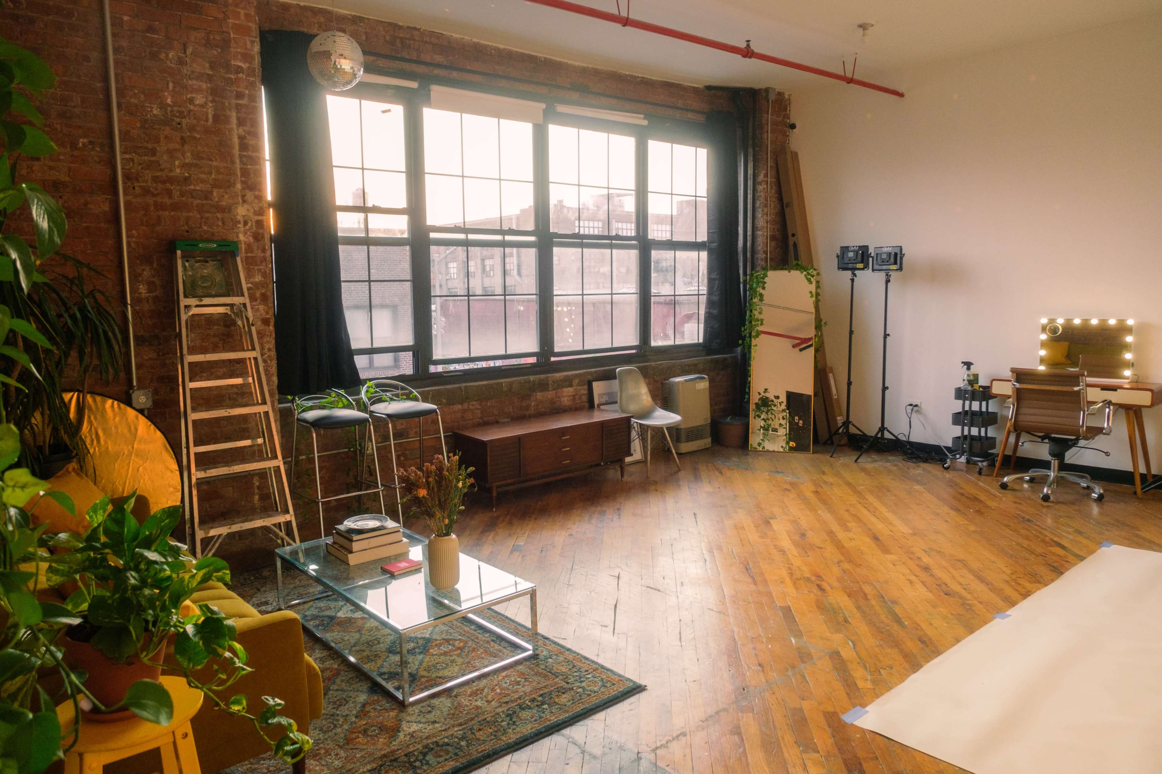 The image depicts a spacious, industrial-style studio with large windows, wooden floors, and various furnishings, including a glass coffee table, bar stools, and a desk with lighting equipment.