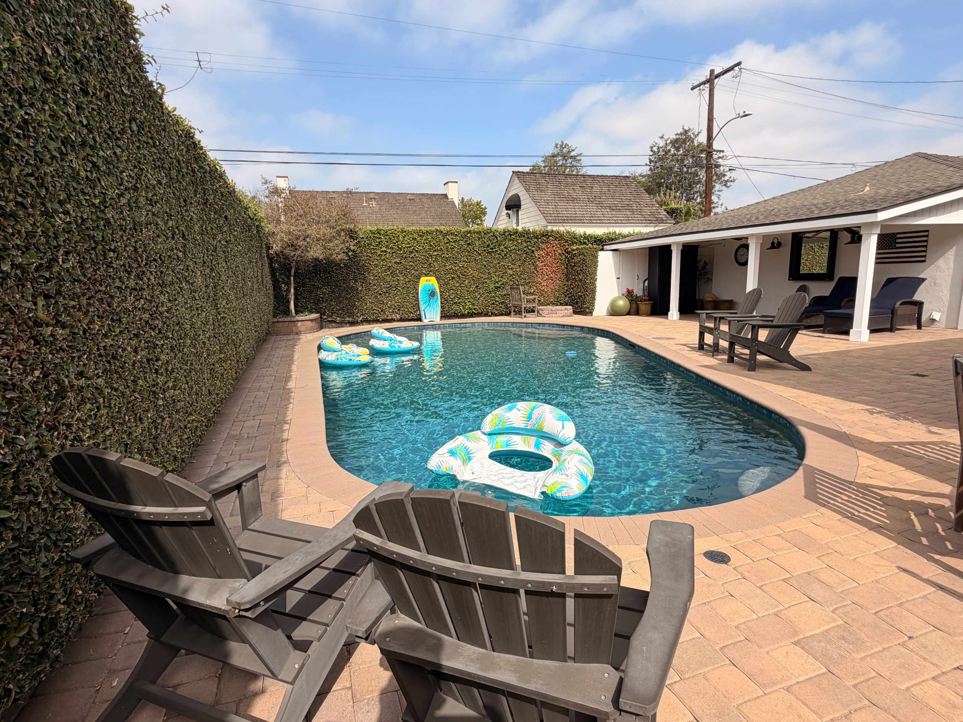 A swimming pool surrounded by hedges, featuring colorful inflatable floats and wooden lounge chairs on a paved patio.