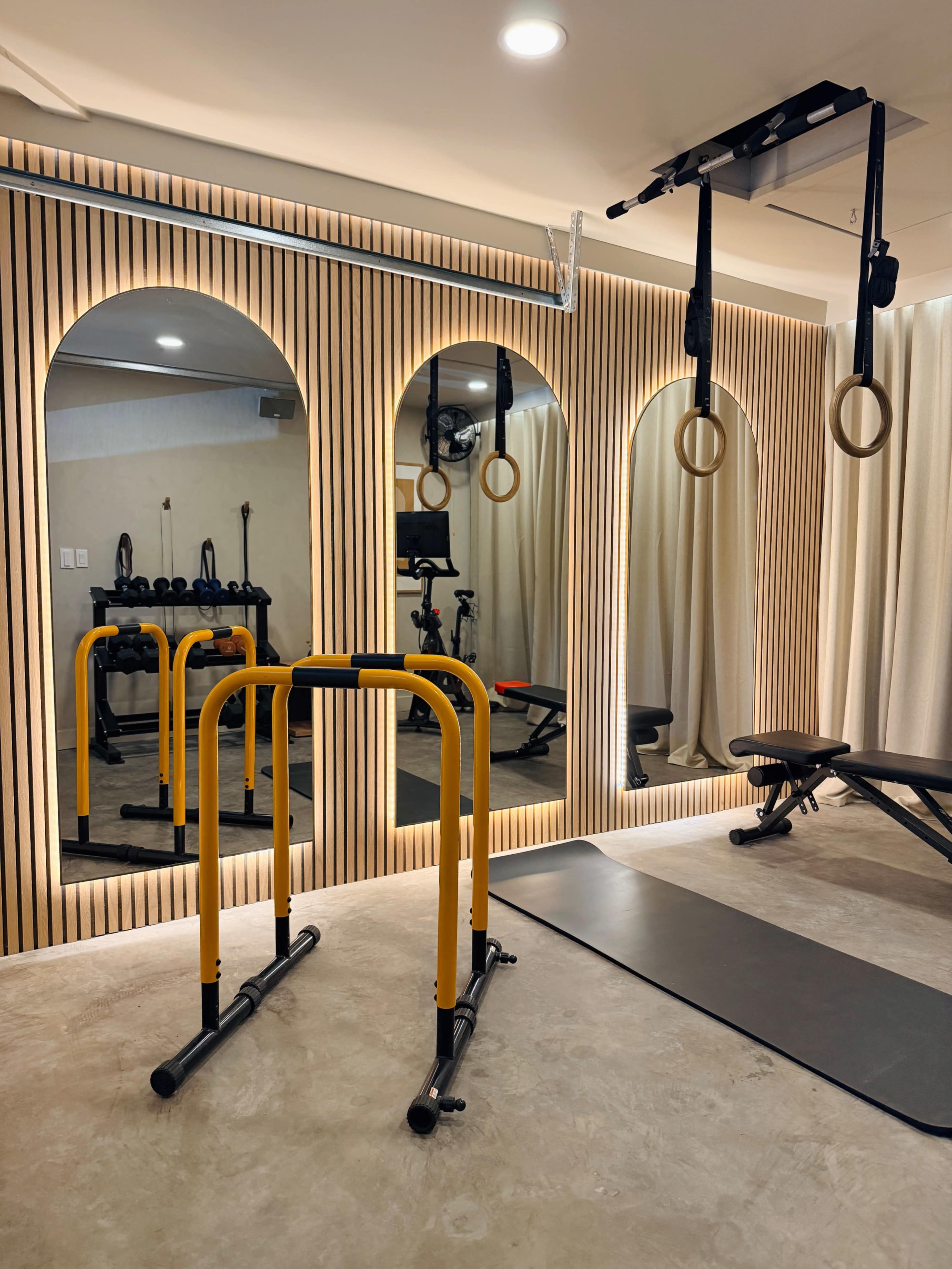 The image shows a modern home gym featuring a set of parallel bars, a weight bench, exercise rings, and a mirrored wall with wooden paneling.