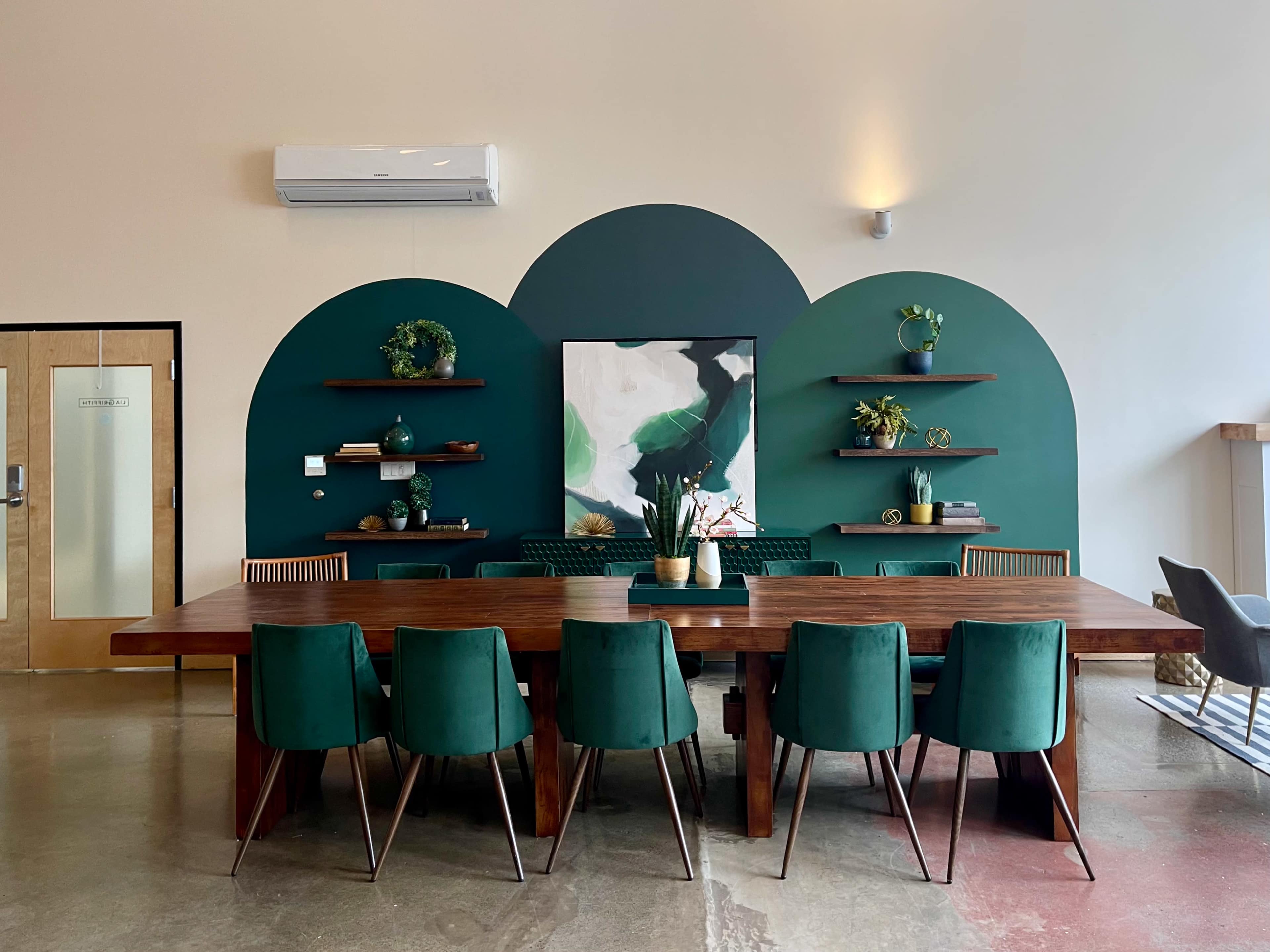 The image shows a modern dining area featuring a large wooden table surrounded by green chairs, with a shaded green wall and shelves displaying plants and artwork in the background.