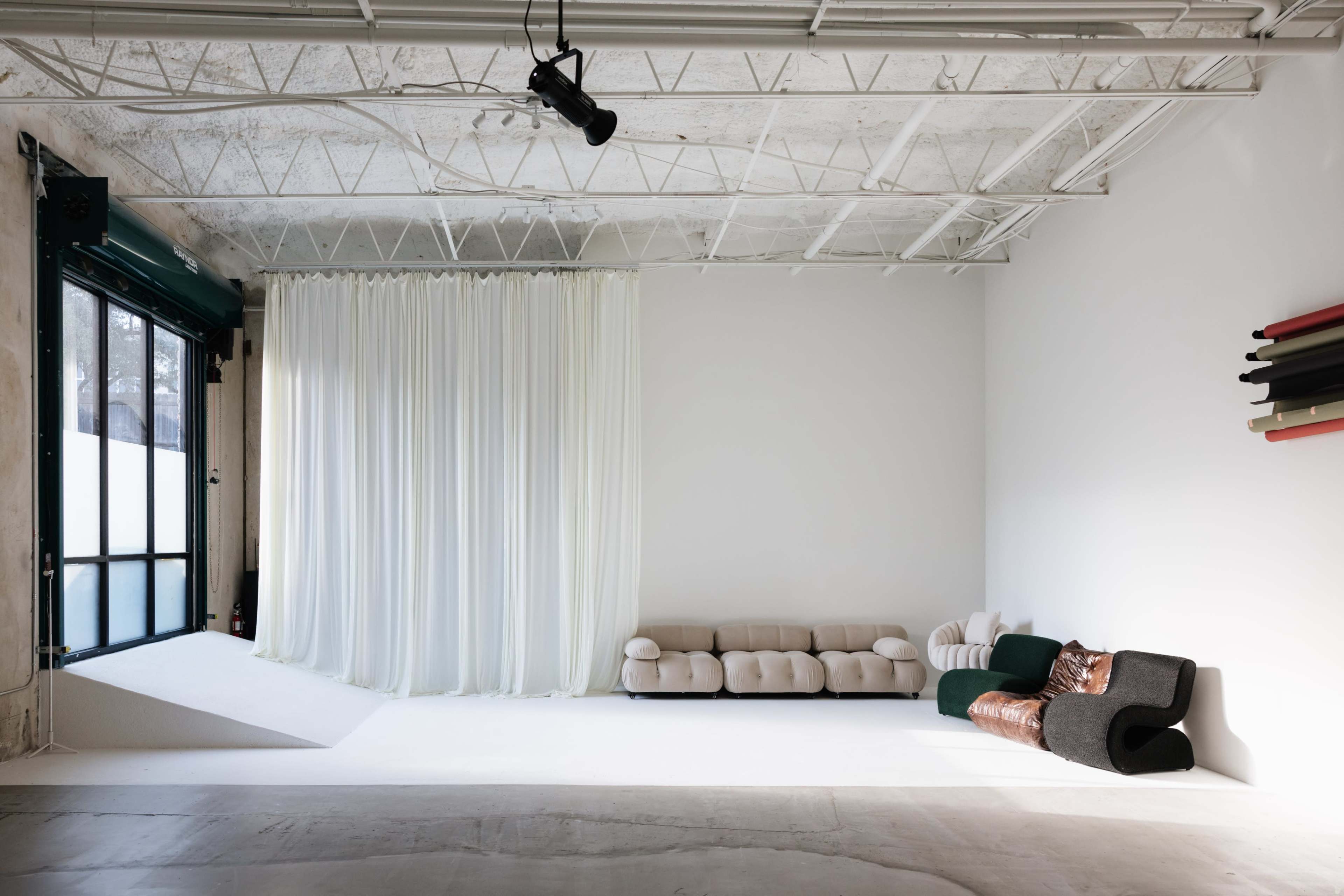 The image shows a spacious, minimalist interior with large windows, soft white curtains, and a combination of modern couches in neutral tones against a plain wall.
