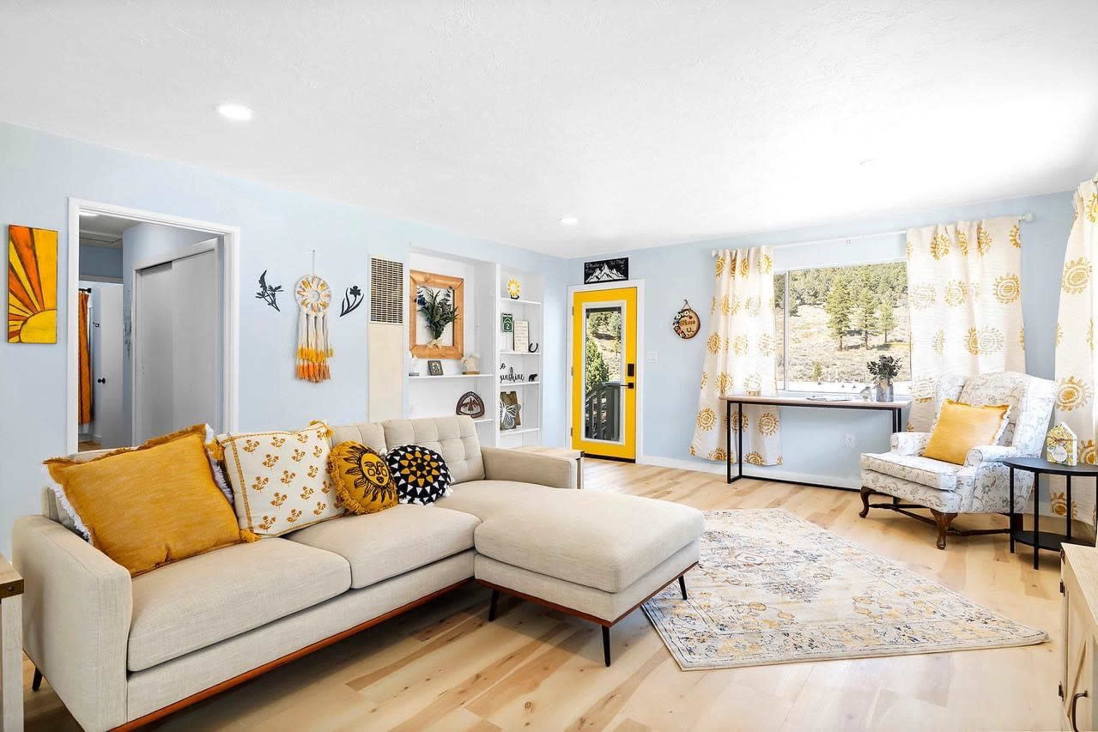 A bright and airy living room features a light-colored sofa with yellow and white accents, a large window with floral curtains, and a cheerful yellow door.