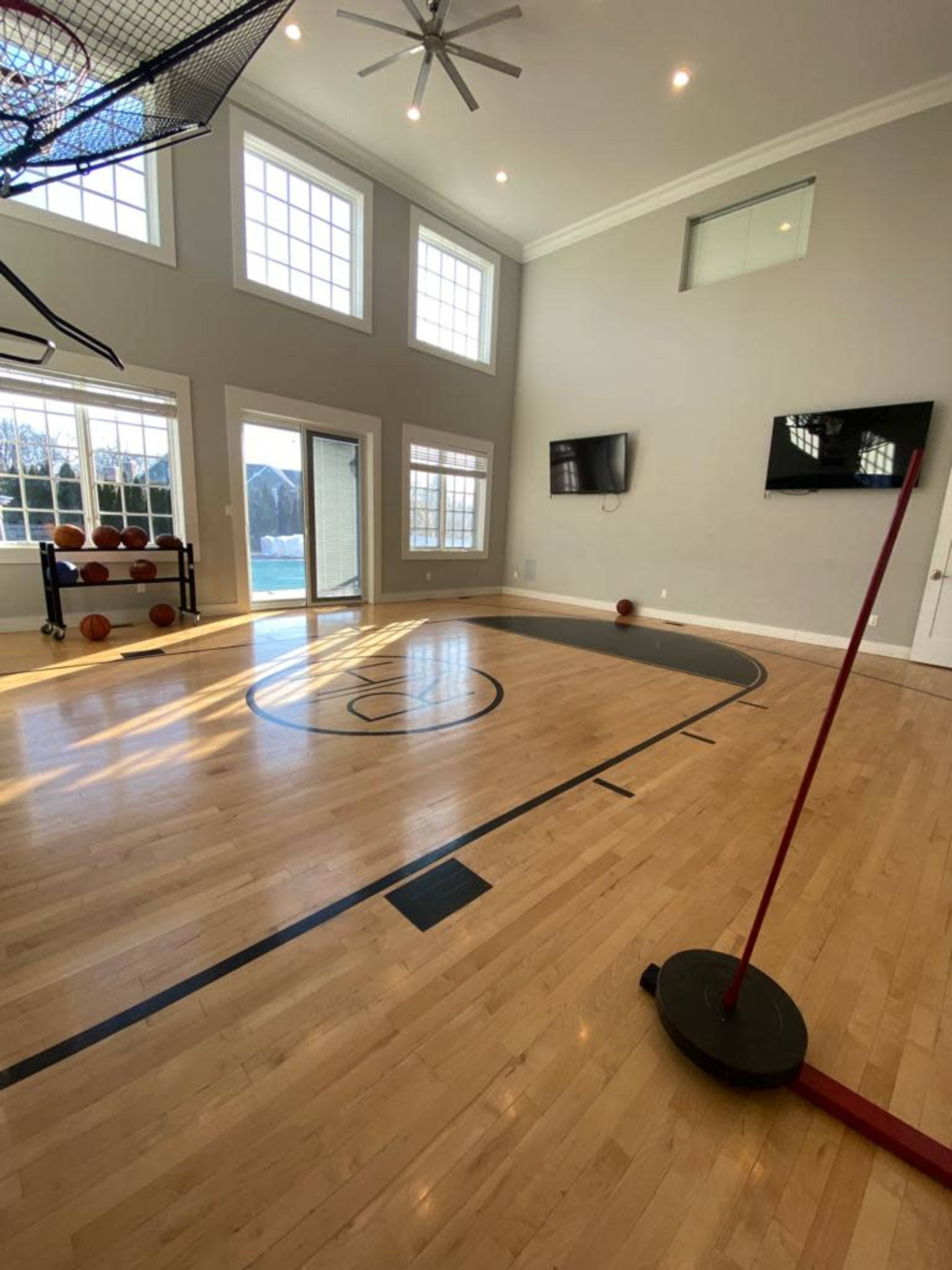 The image shows an indoor basketball court with a hoop, several basketballs on the floor, and large windows allowing natural light.