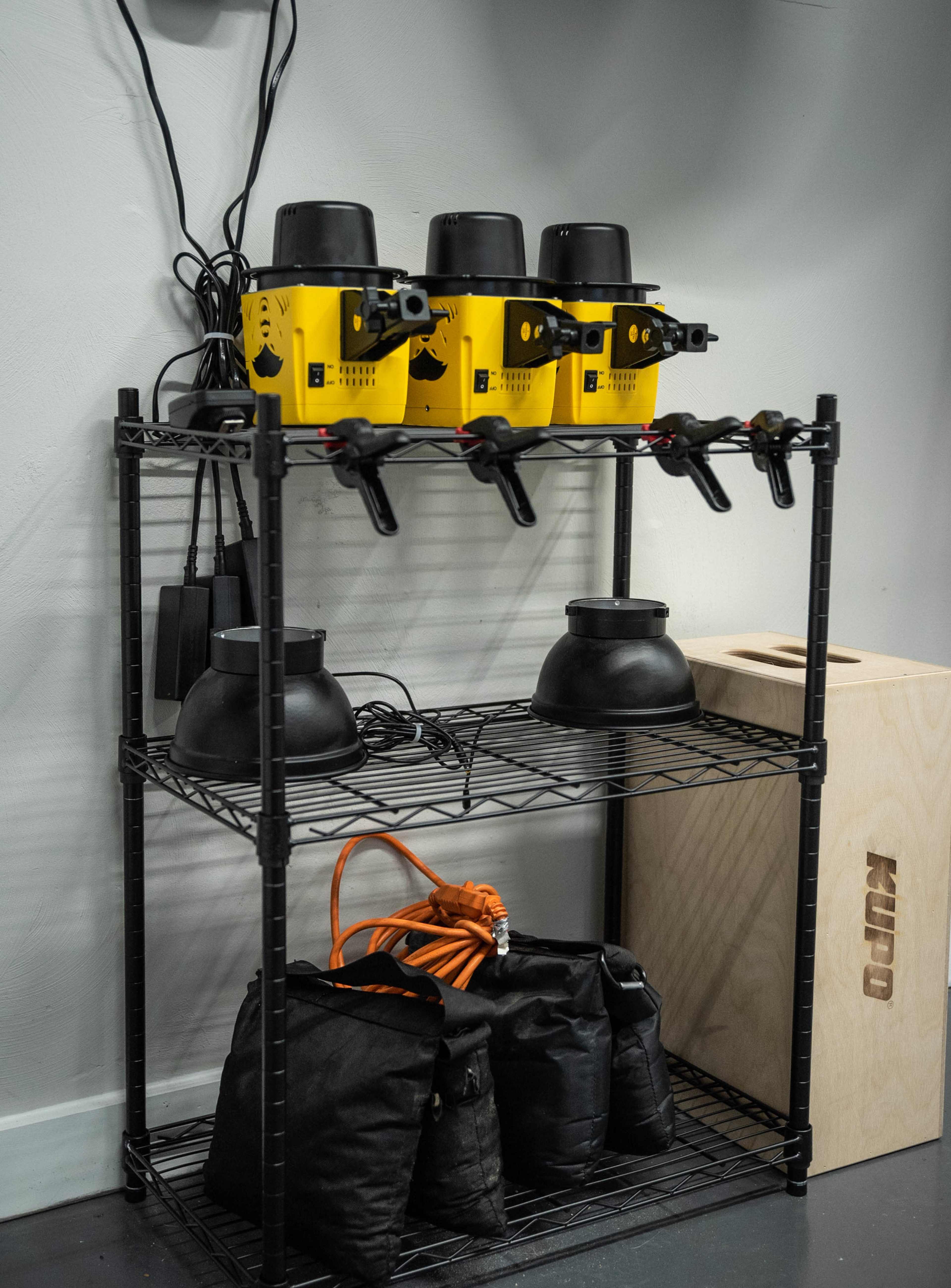 A wire shelving unit with three yellow machines on the top shelf, two black bowls and an orange extension cord on the middle shelf, and black bags on the bottom shelf.