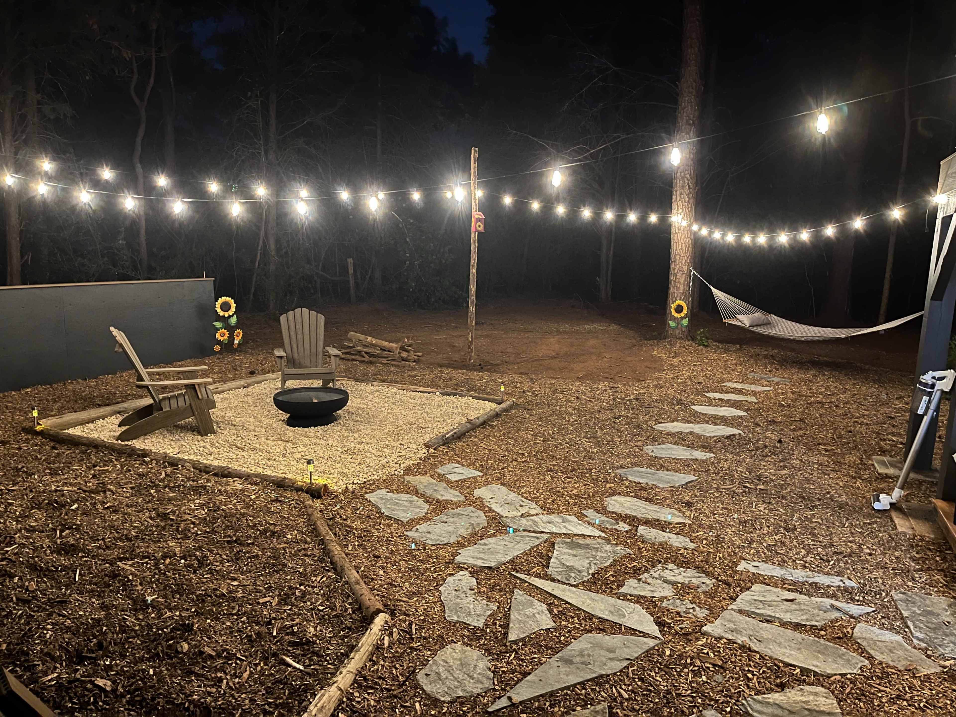 The image shows a cozy outdoor area with a gravel seating space, a fire pit, a stone path, and string lights hanging in the trees, along with a hammock nearby.