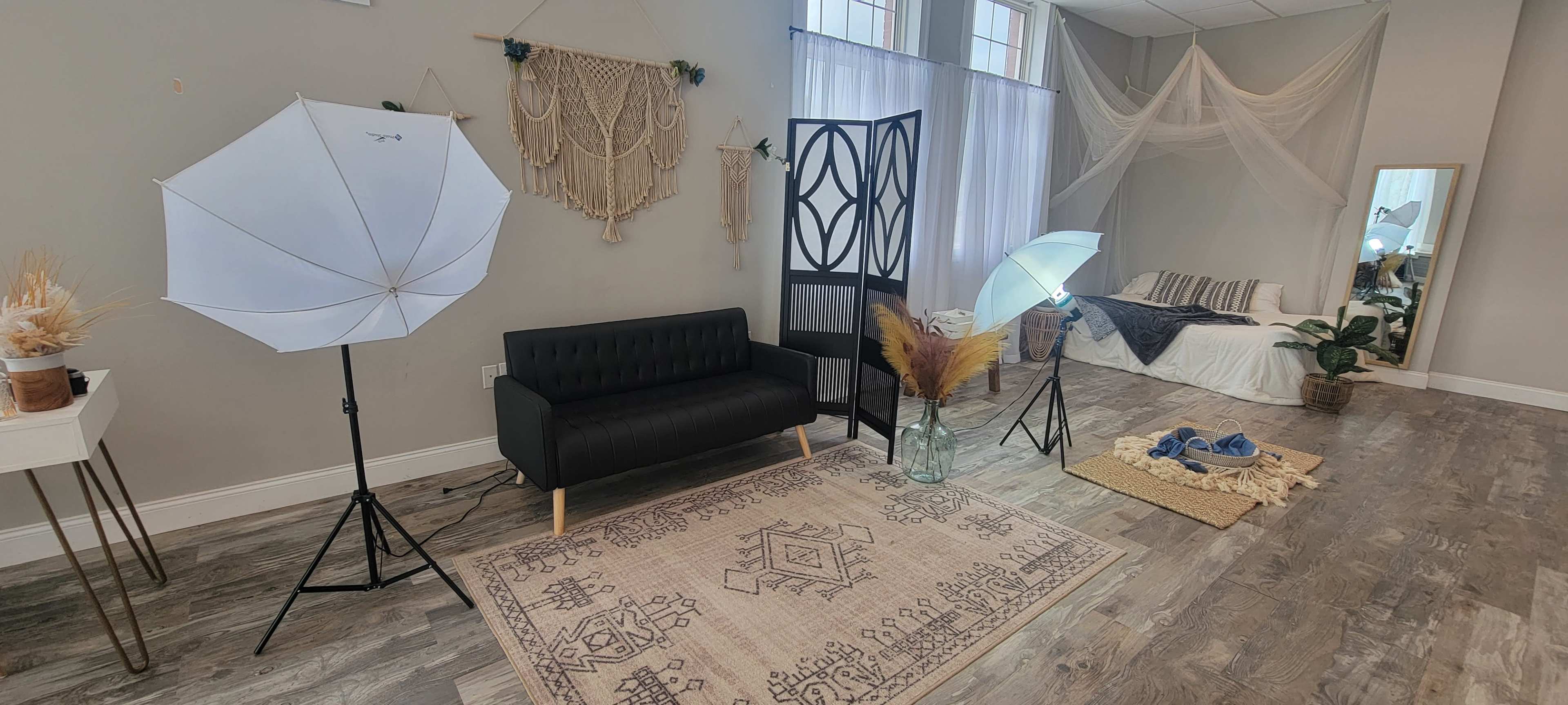 A photography studio with a black couch, two lighting umbrellas, a decorative backdrop, and a cozy seating area with a bed and various textiles.