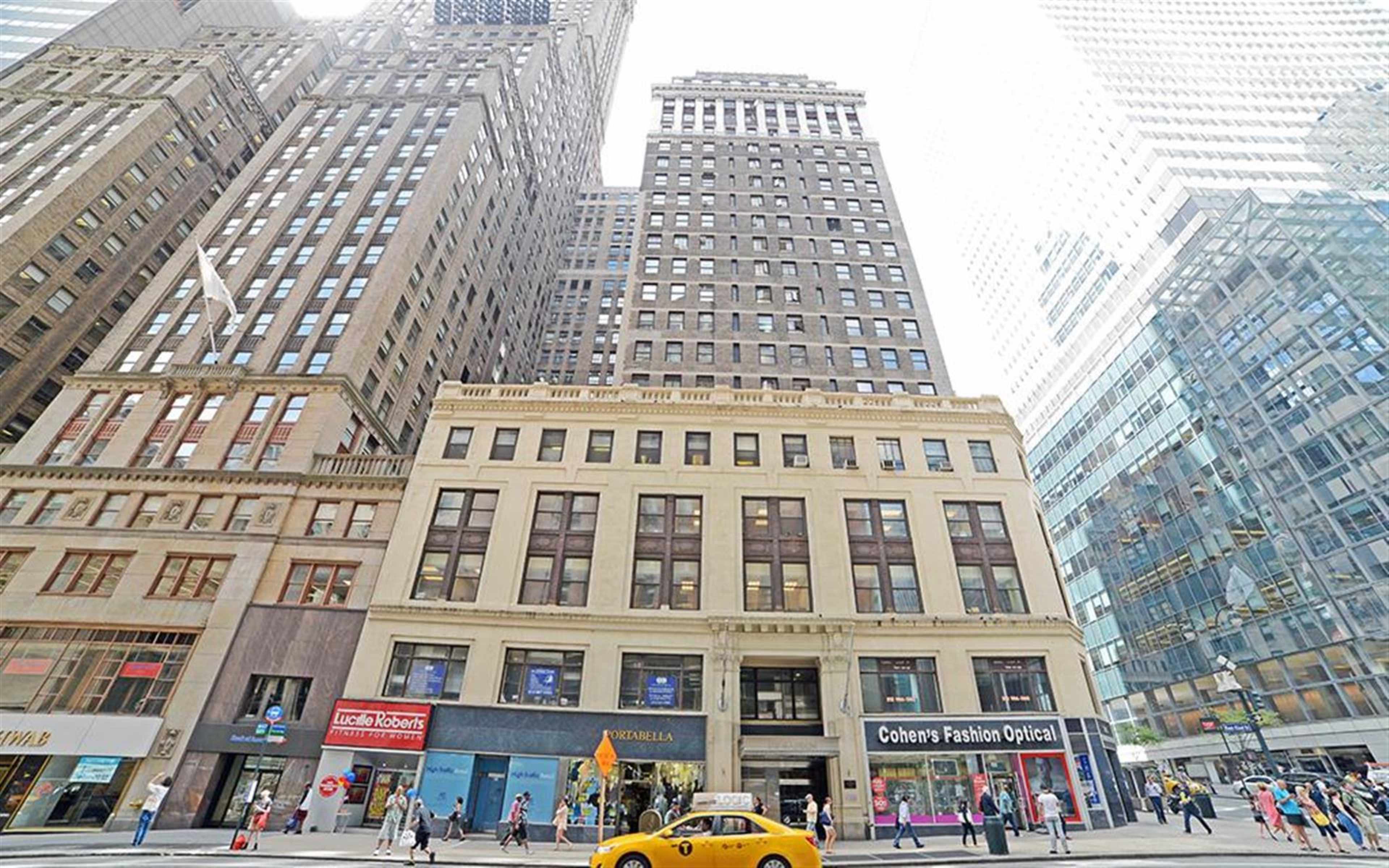 A street view of tall buildings in a bustling urban environment, with a yellow taxi parked in front of a row of storefronts.