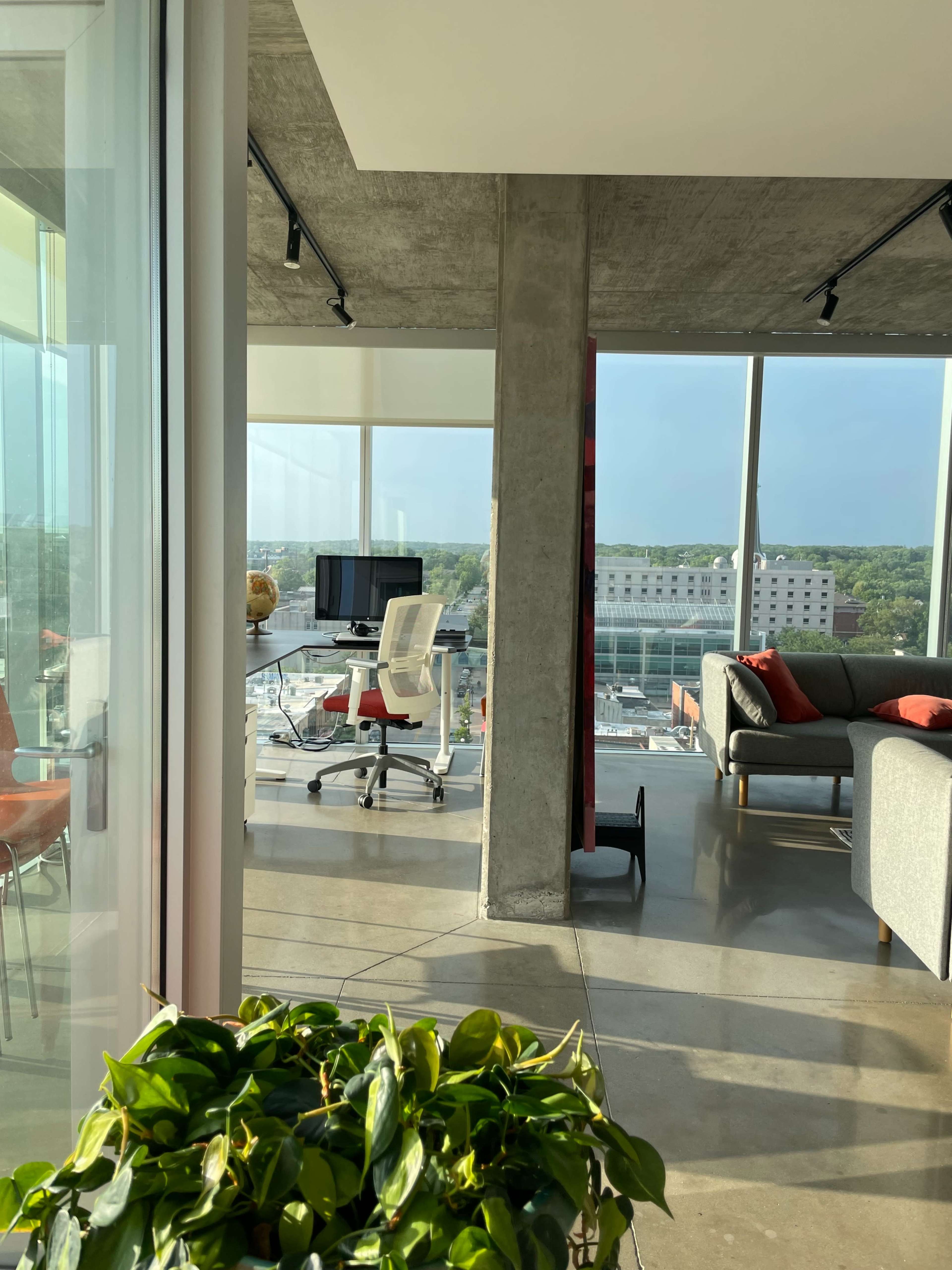 A modern office space featuring a desk with a computer, a comfortable chair, and a sofa, all set against large windows that offer a view of the city.