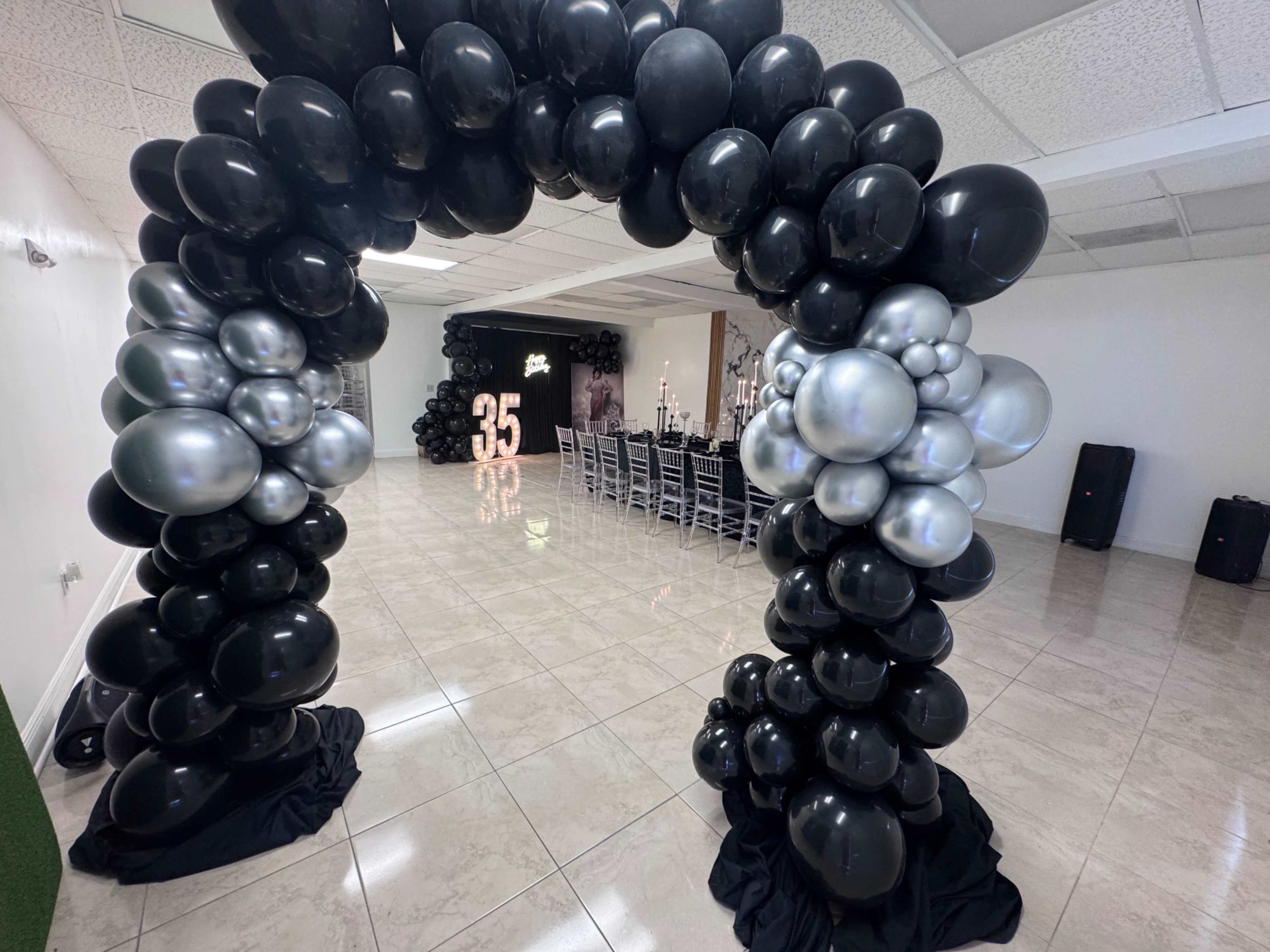 The image shows a room decorated with a large black and silver balloon arch, leading to a dining area set with tables and illuminated number "35" in the background.